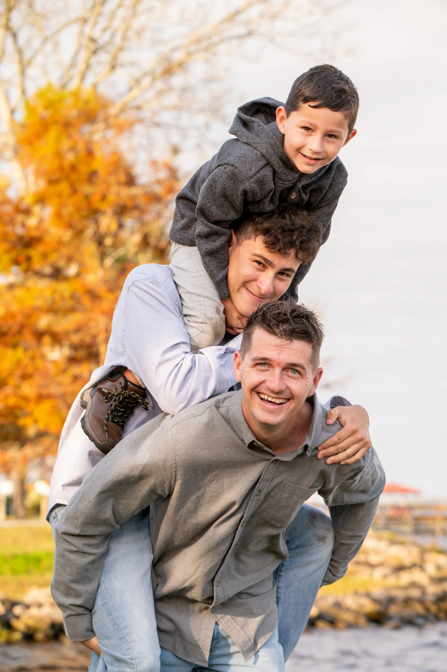 A man with two boys on his back, outdoors in a park with autumn trees in the background. They are smiling and enjoying each other's company during fall.