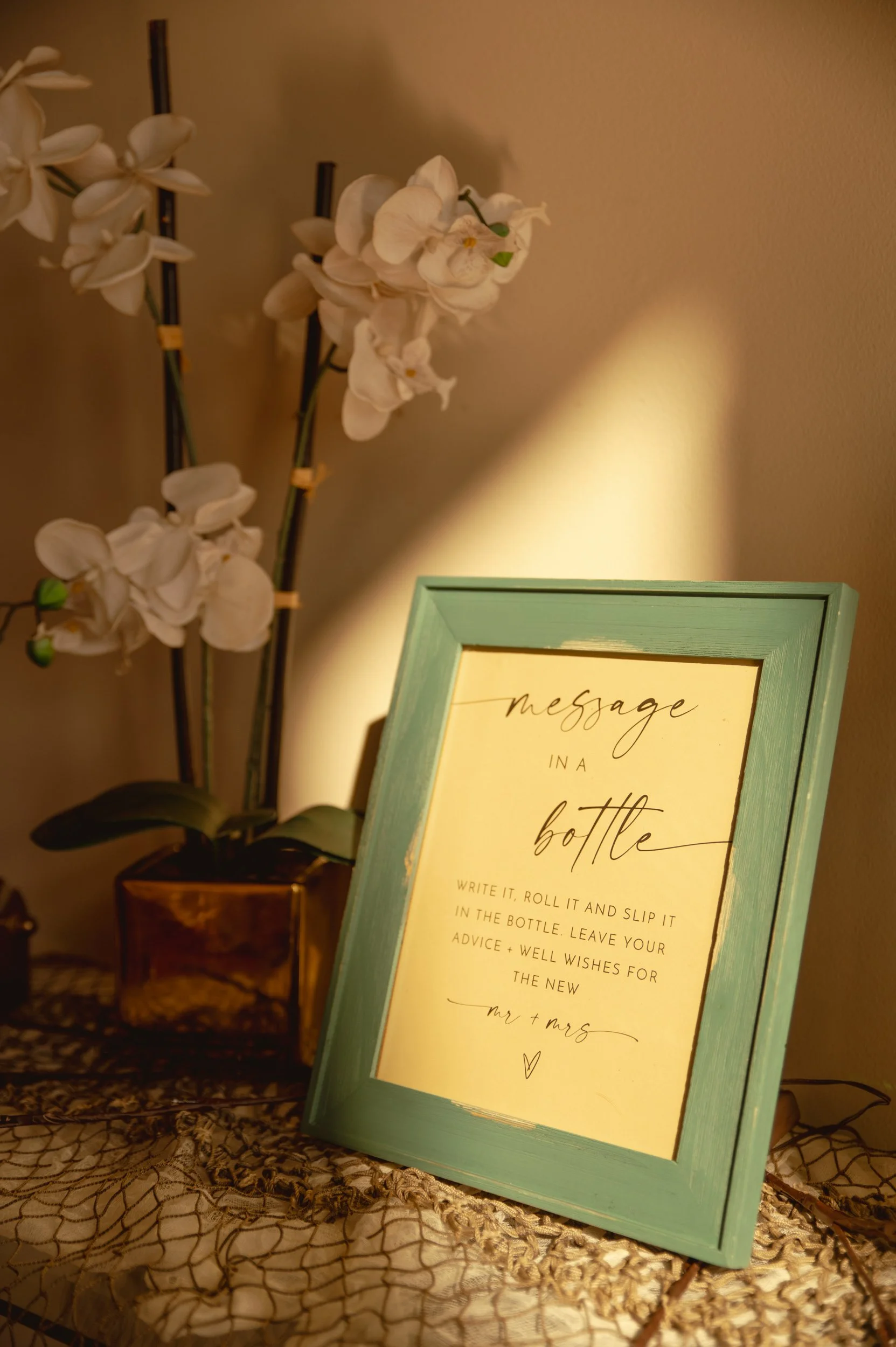 A framed sign with the text 'message in a bottle' on a table next to a potted orchid with white flowers.