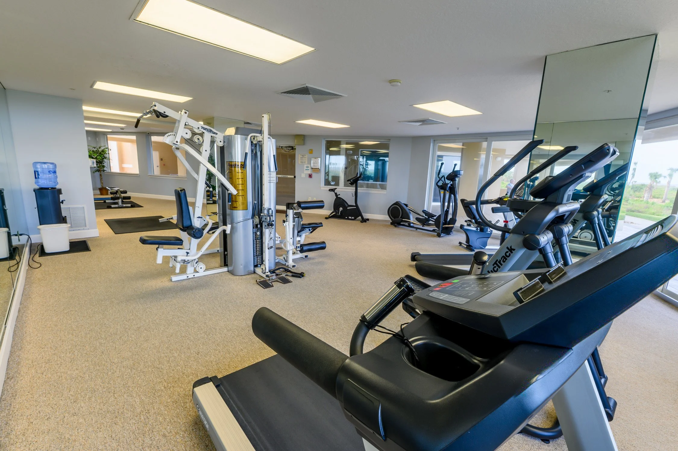 Empty gym room with cardio and strength training equipment, including treadmills, ellipticals, a water dispenser, and padded flooring.