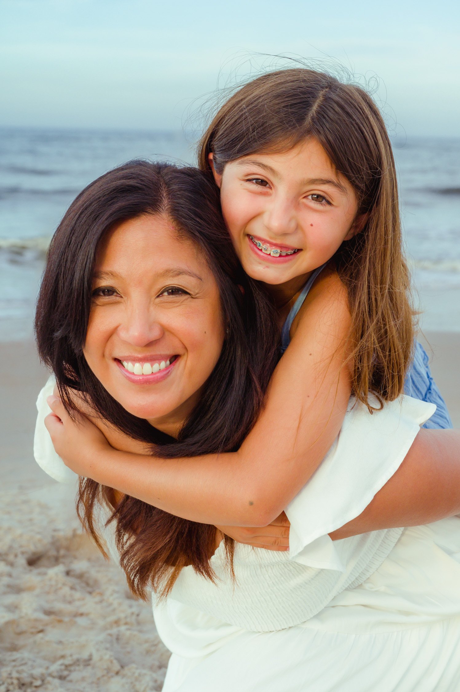 A woman and a girl smiling on a beach, with the girl piggyback riding the woman, both showing happiness.