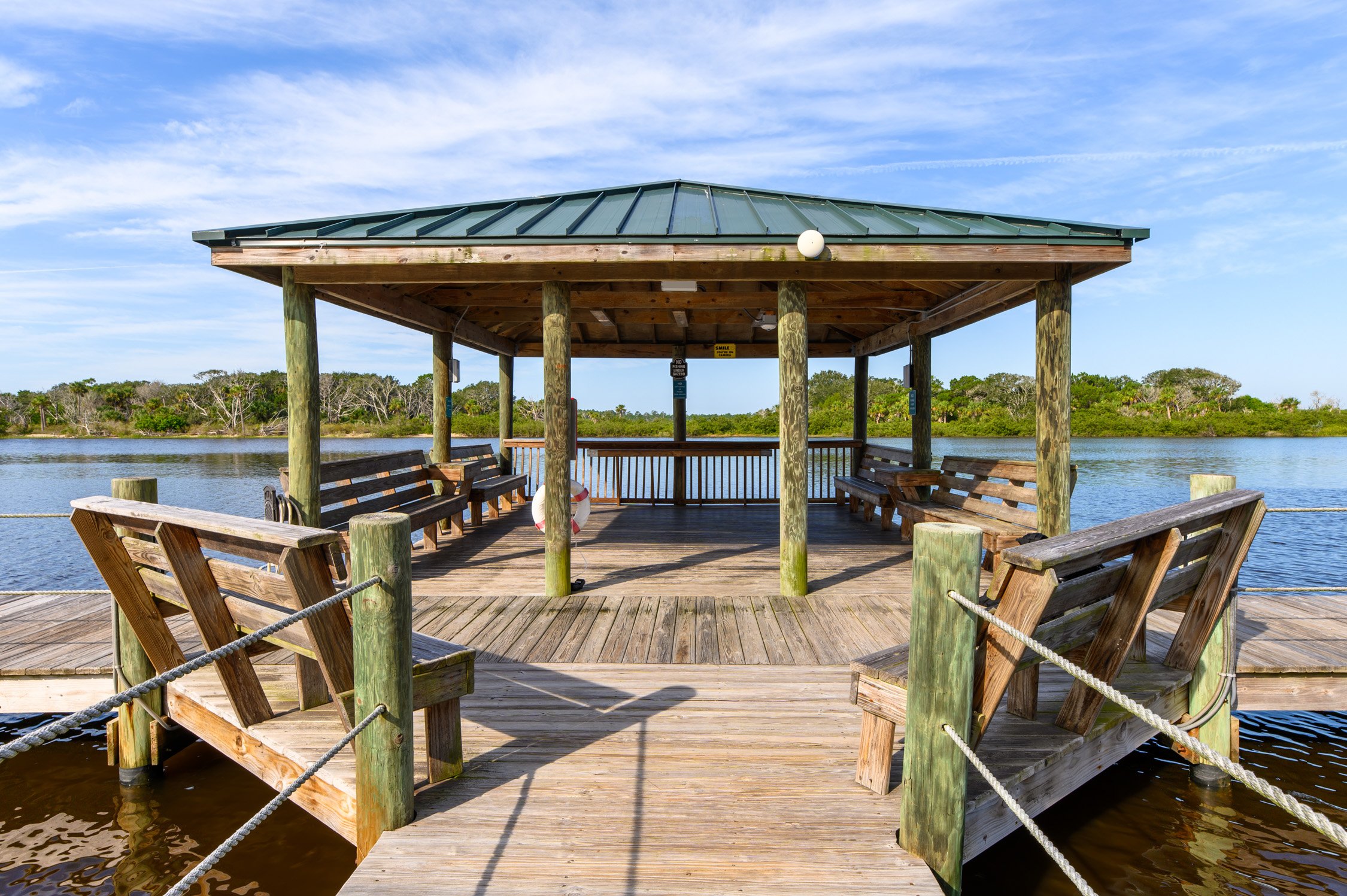 A wooden gazebo with benches on a pier over water, surrounded by lush green trees and a blue sky.