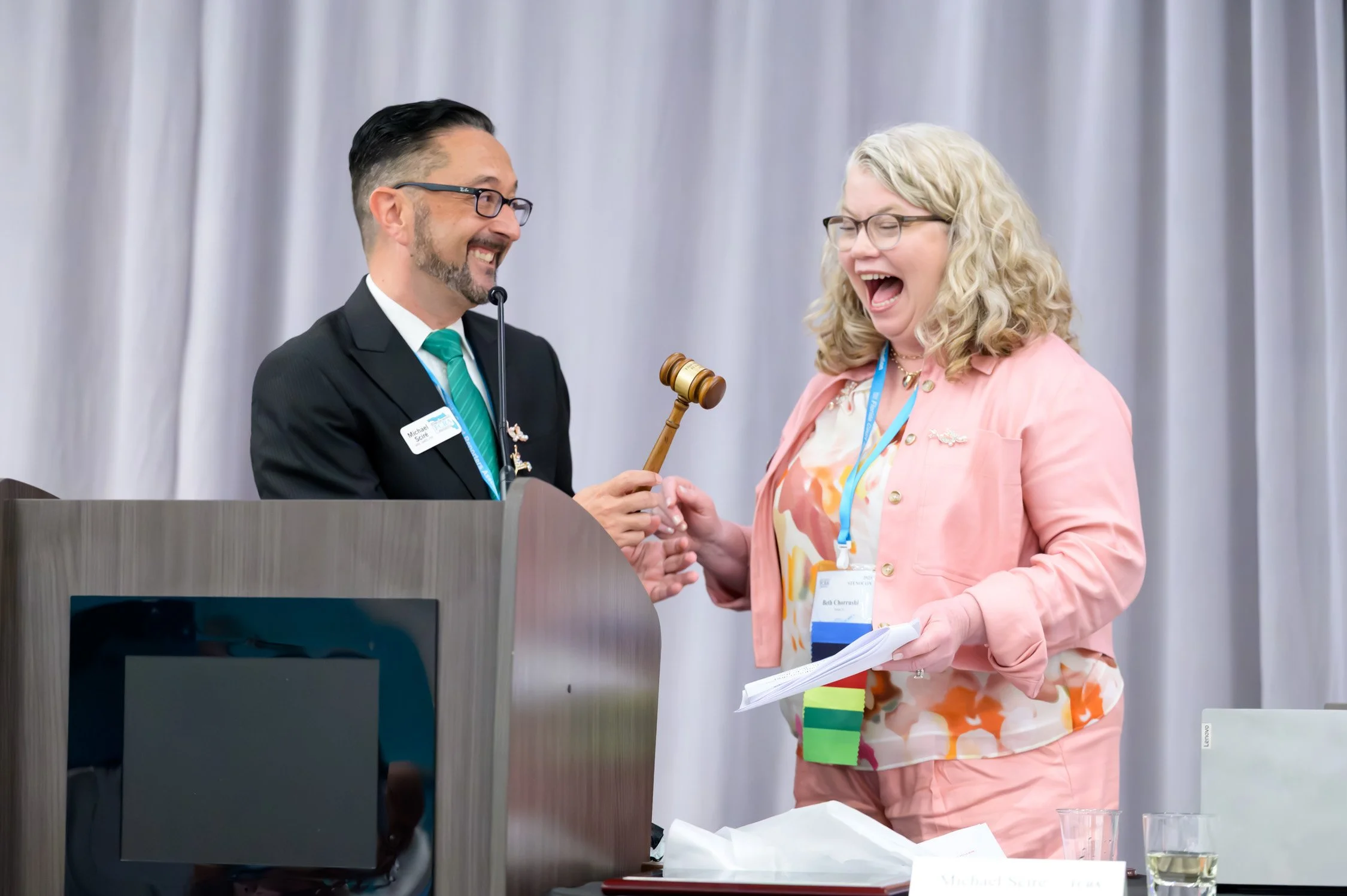 A man in a suit and woman in pink attire are on stage. The man is holding a gavel, and both are smiling and laughing. There are papers and glasses on the table in front of them.