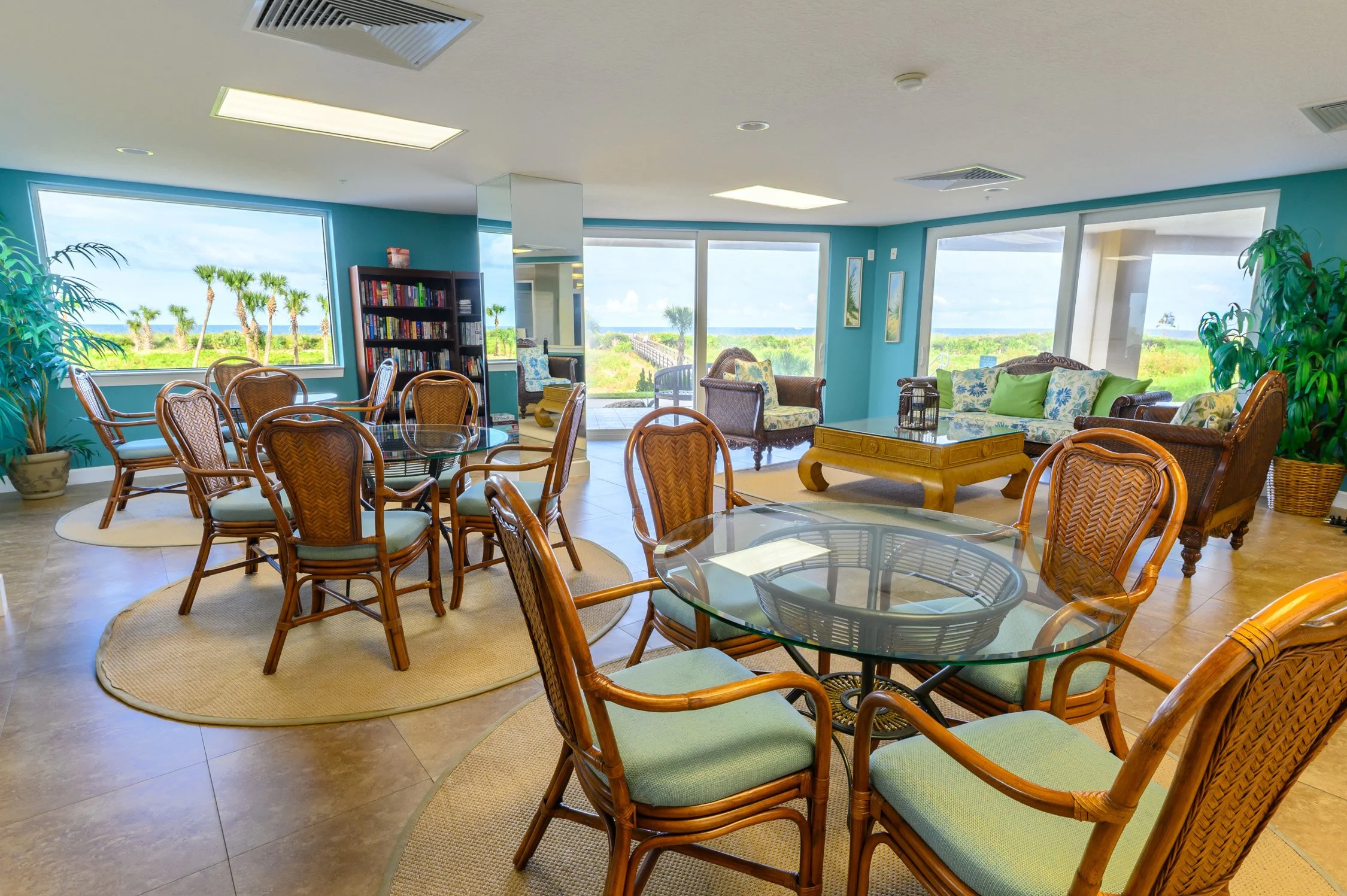A bright, spacious indoor lounge area with large windows showcasing a view of palm trees and greenery outside. The room features multiple round and rectangular glass tables with wooden chairs that have light green cushions. There is a bookshelf with 