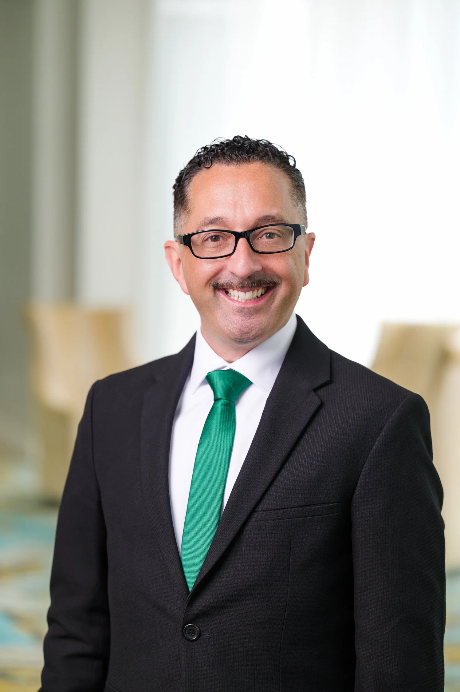 A smiling man wearing a black suit, white shirt, green tie, and glasses, standing indoors with a blurred background.