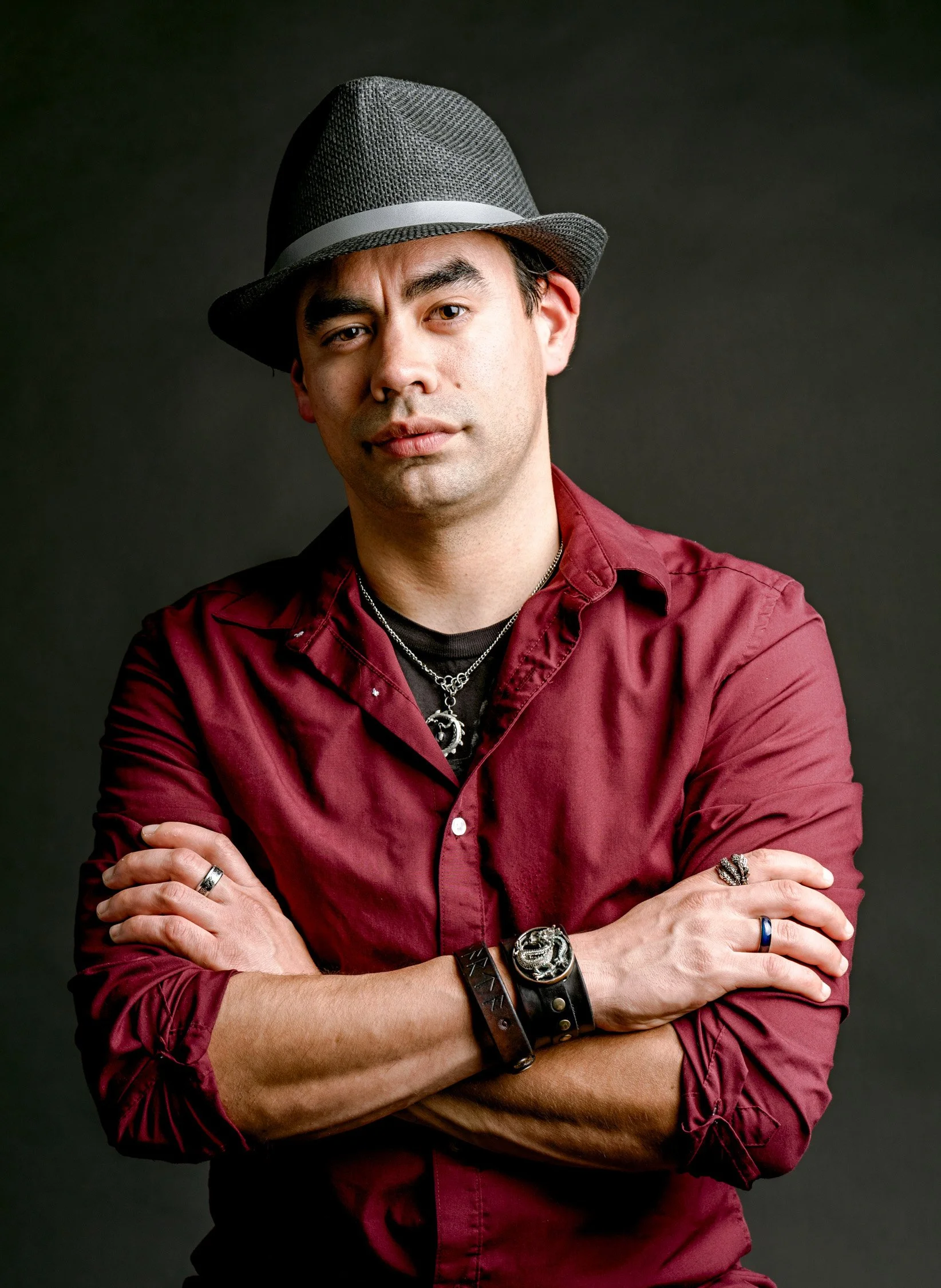 A man with light skin wearing a gray fedora hat, a red button-up shirt with sleeves rolled up, multiple rings, bracelets, necklaces, and styled hair, standing with arms crossed against a dark background.
