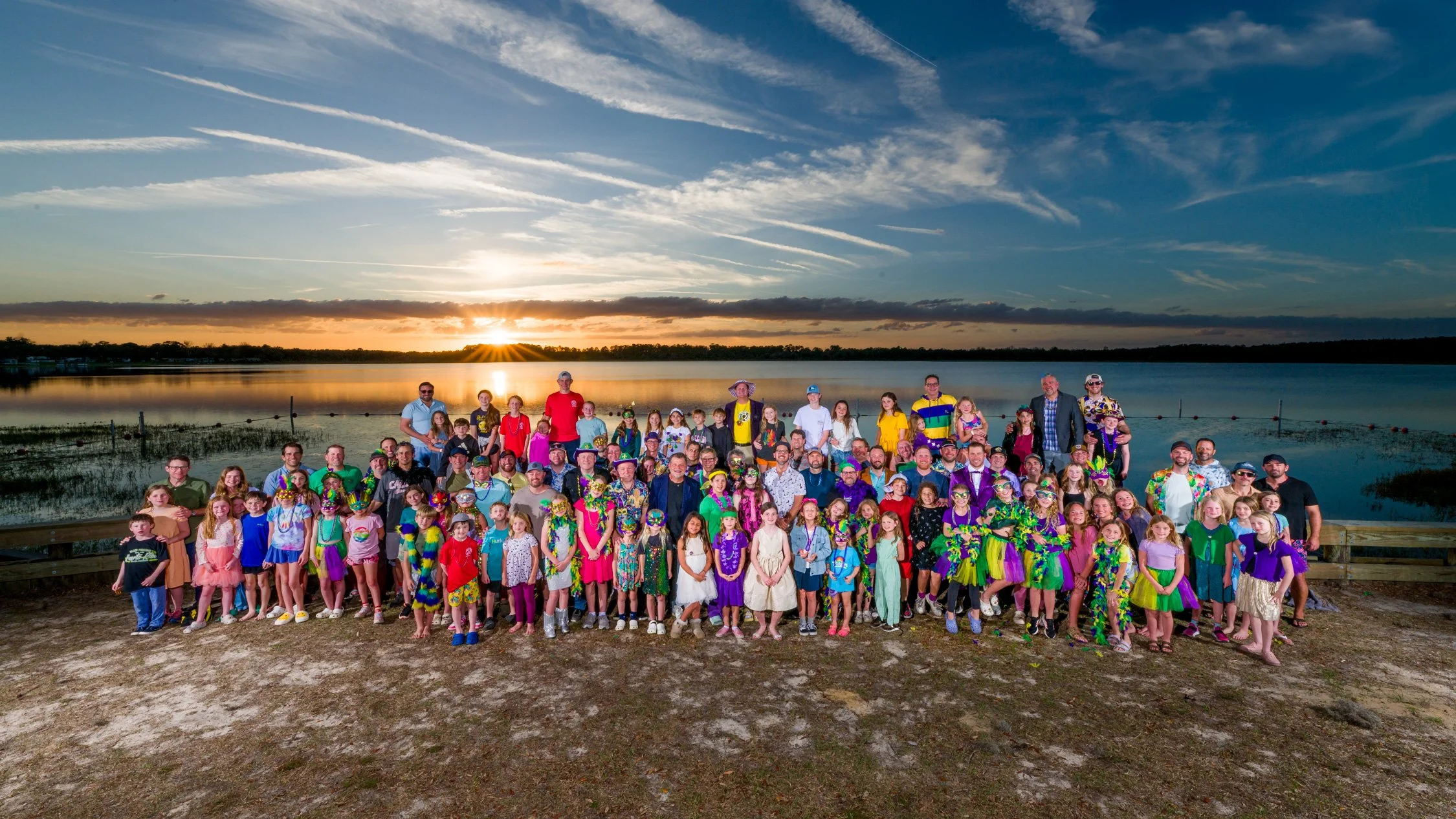A large group of people, including children and adults, standing together outdoors near a body of water during sunset. Many are dressed in colorful, festive costumes and outfits.