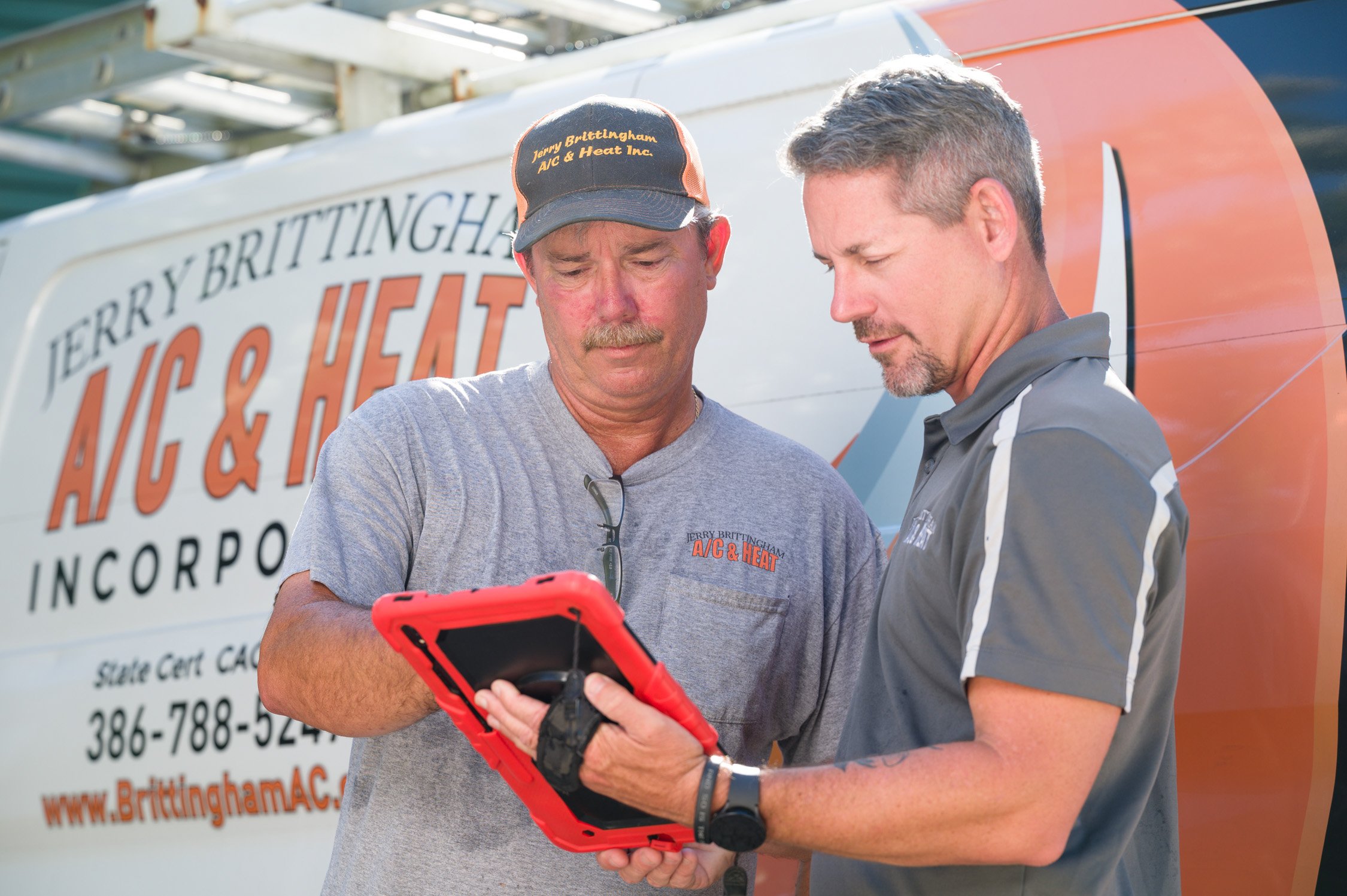 Two men stand outdoors in front of a van with the logo 'Terry Brittingham A/C & Heat Inc.' on it. The man on the left, wearing a gray t-shirt and a cap, looks at a digital device held by the man on the right, who is wearing a gray polo shirt. They ap