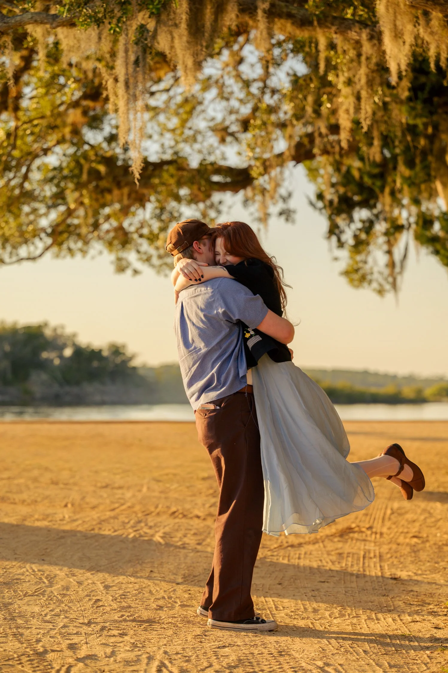 A couple embracing and smiling outdoors during sunset, with the woman lifted off the ground. They are standing on a sandy area near a body of water, with large hanging moss and trees in the background.