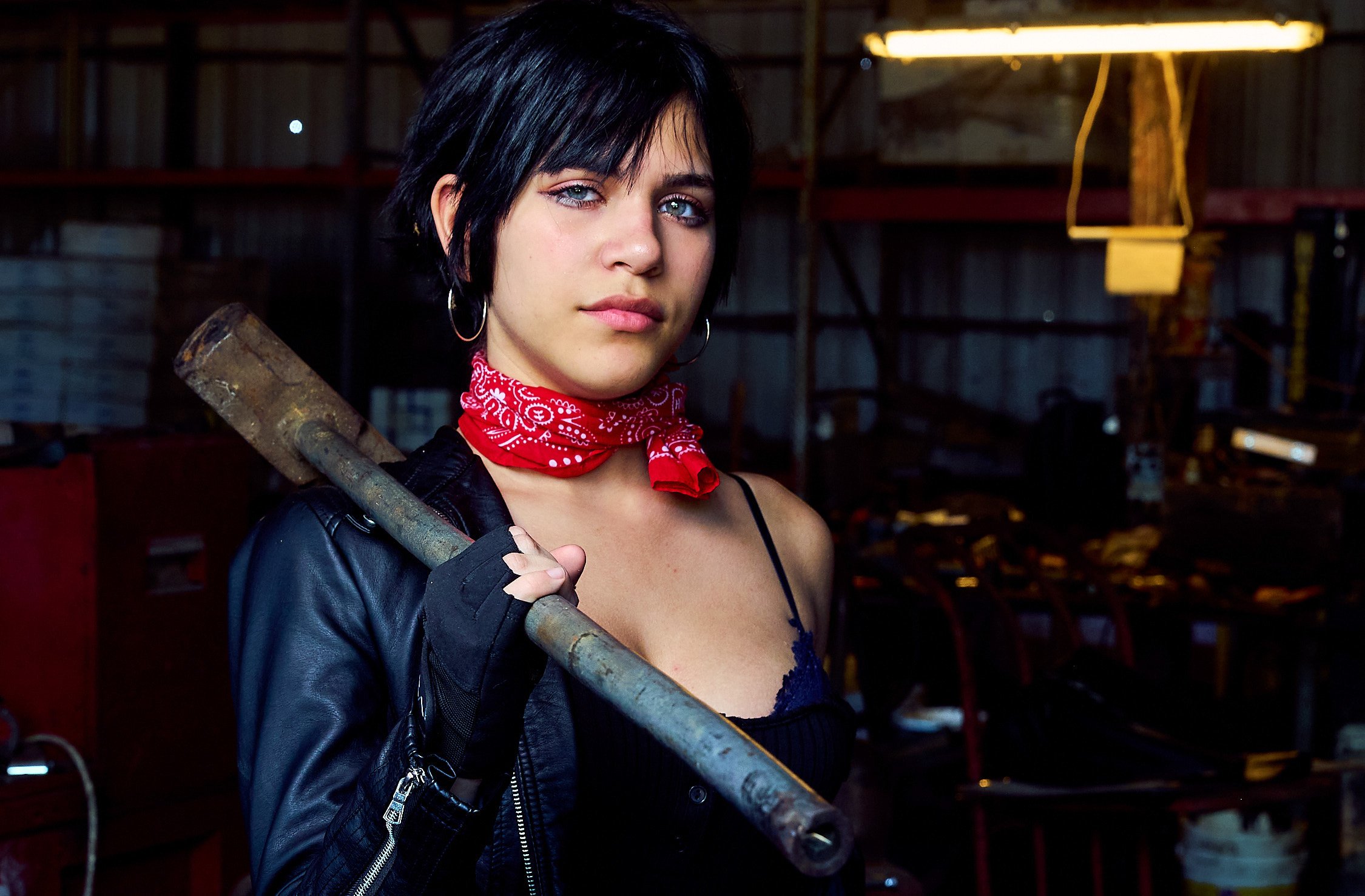 A woman with black hair, blue eyes, and hoop earrings in a workshop, holding a rusty sledgehammer over her shoulder, wearing a red bandana around her neck, a black leather jacket, and a black top.