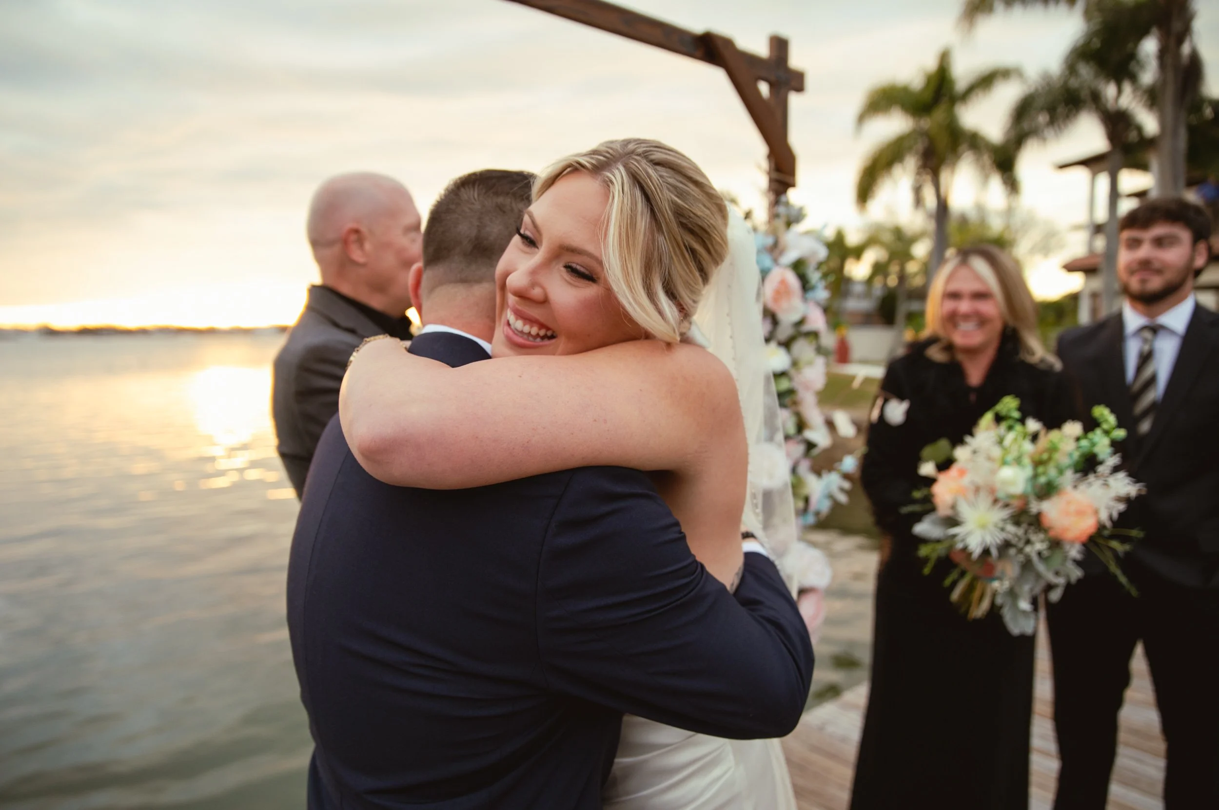 A bride and groom embrace during their wedding ceremony at sunset near a body of water, with guests and floral arrangements in the background.