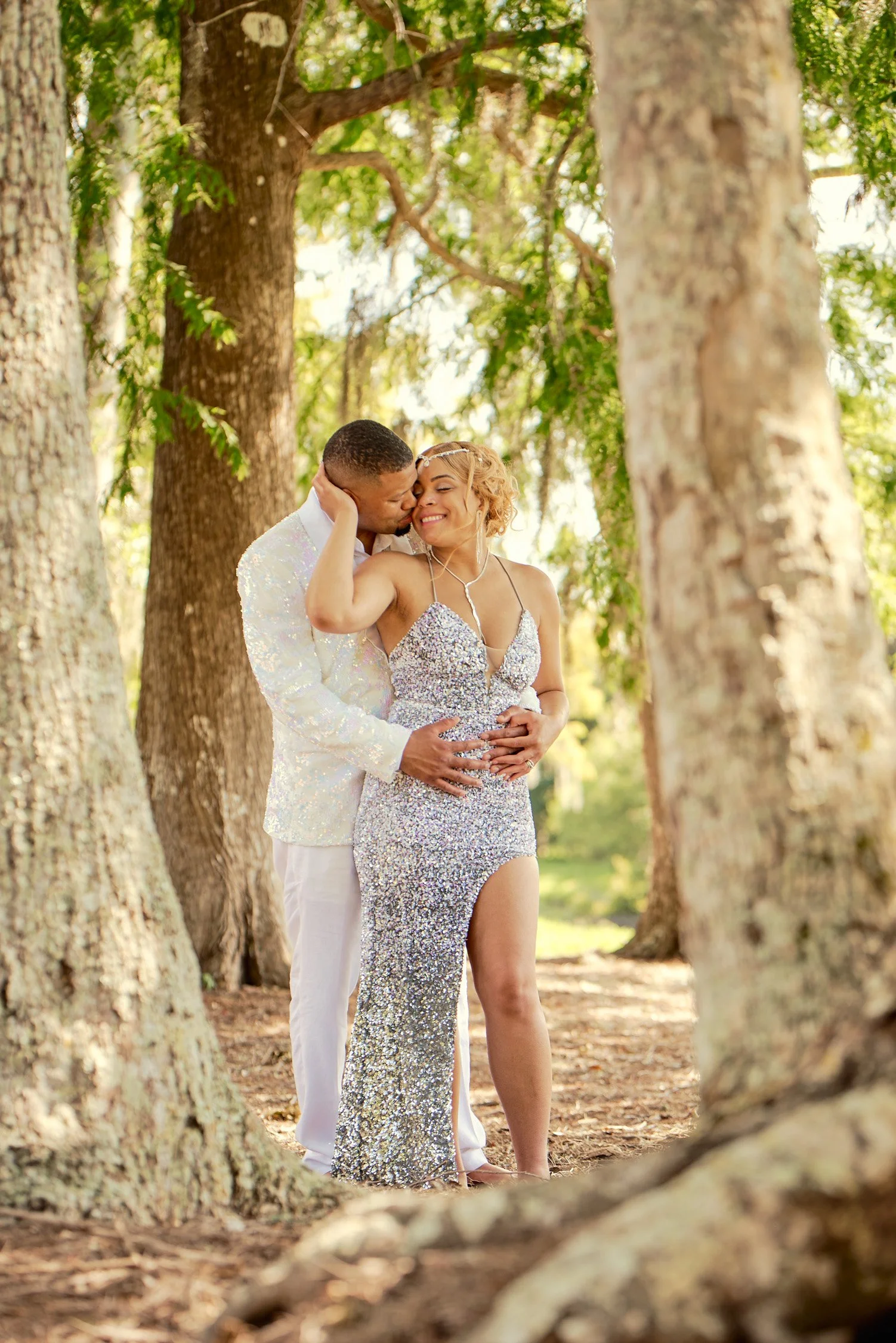 A couple in formal wear sharing an intimate moment in a wooded area during daytime. The woman wears a sparkling silver gown and the man wears a white suit, both smiling and embracing.