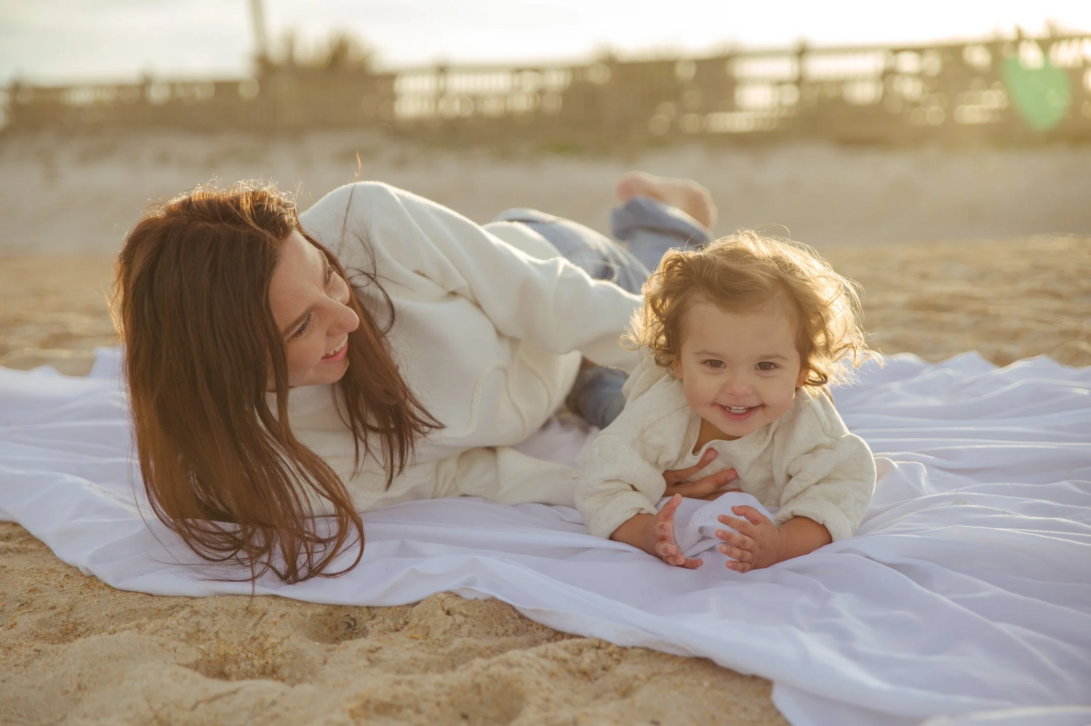 A woman and a small child play on a beach with a white blanket, smiling and enjoying the sunny weather.