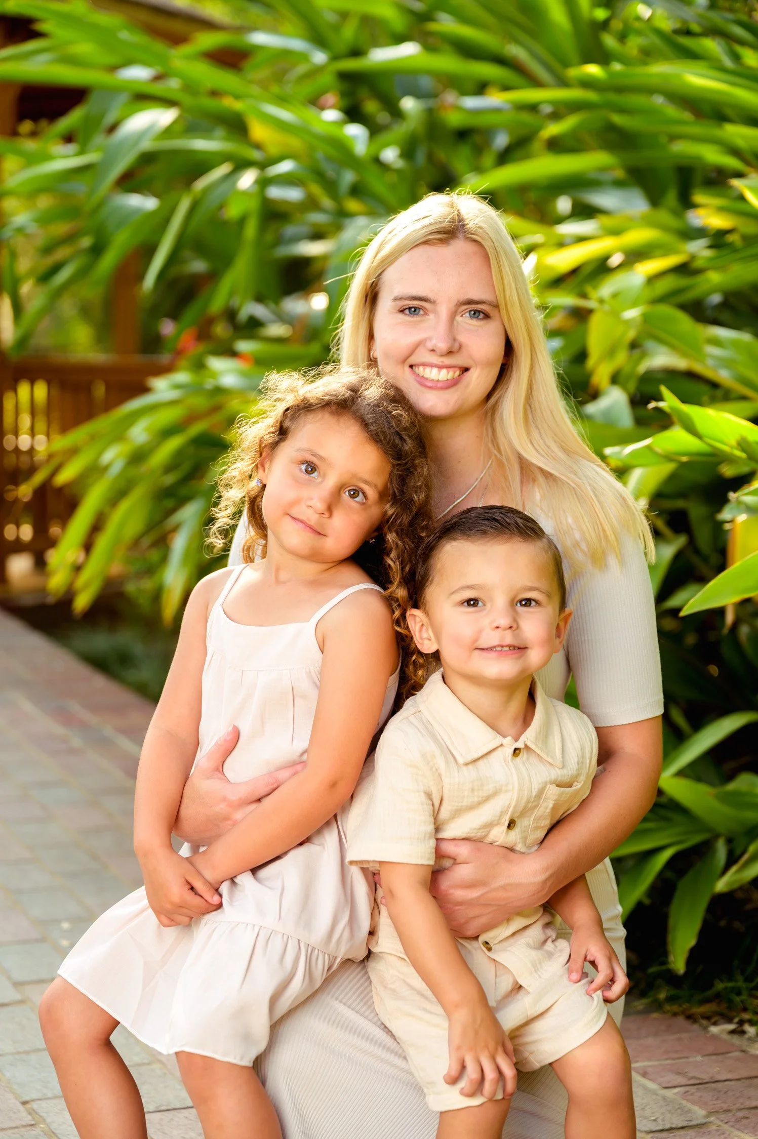 A smiling woman with long blonde hair is sitting outdoors with two young children, a girl and a boy, surrounded by lush green plants, during daylight.