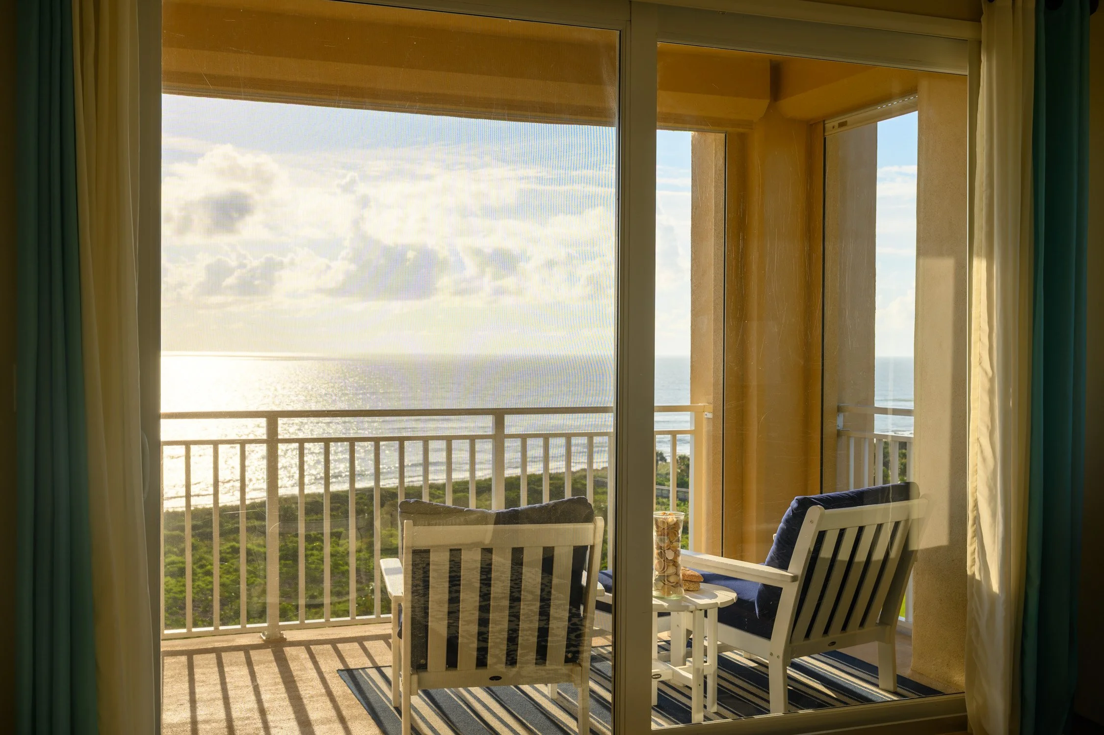 View of a balcony overlooking the ocean with two chairs and a small table, sunlight shining through clouds.