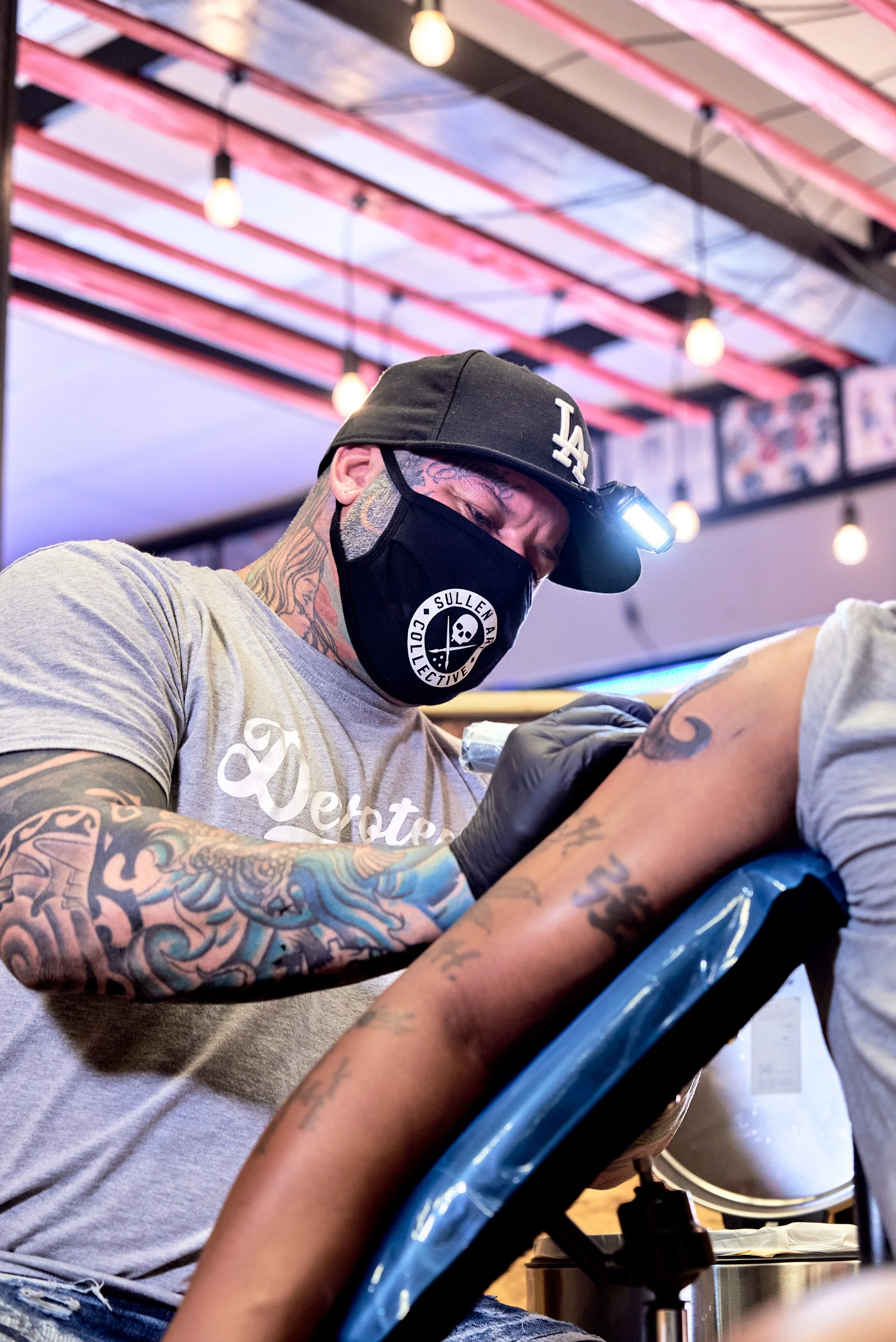 Tattoo artist wearing a black face mask, cap, and gloves, tattooing someone's arm in a tattoo studio with colorful ceiling lights.