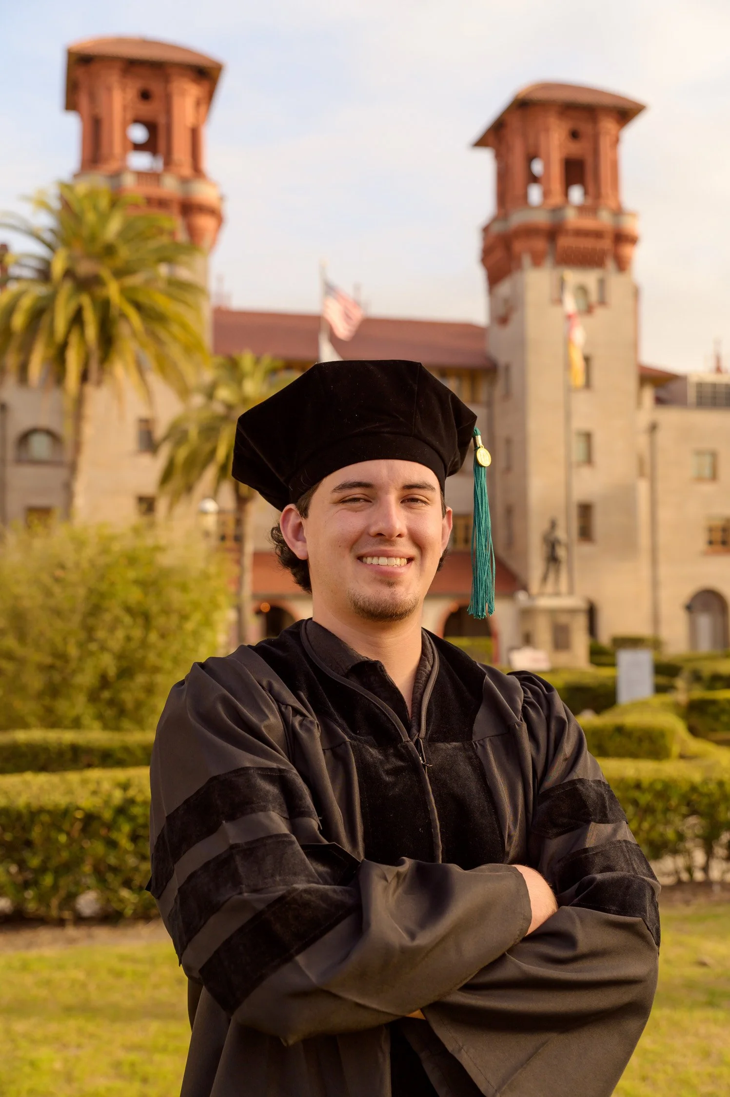 Graduate wearing a cap and gown, standing in front of a historic building with towers, palm trees, and greenery.