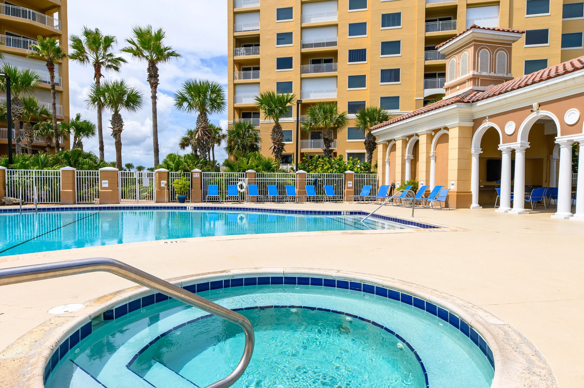 A pool area with a hot tub in the foreground, blue lounge chairs along the pool’s edge, a fence with palm trees behind, and a large yellow apartment building in the background under a partly cloudy sky.
