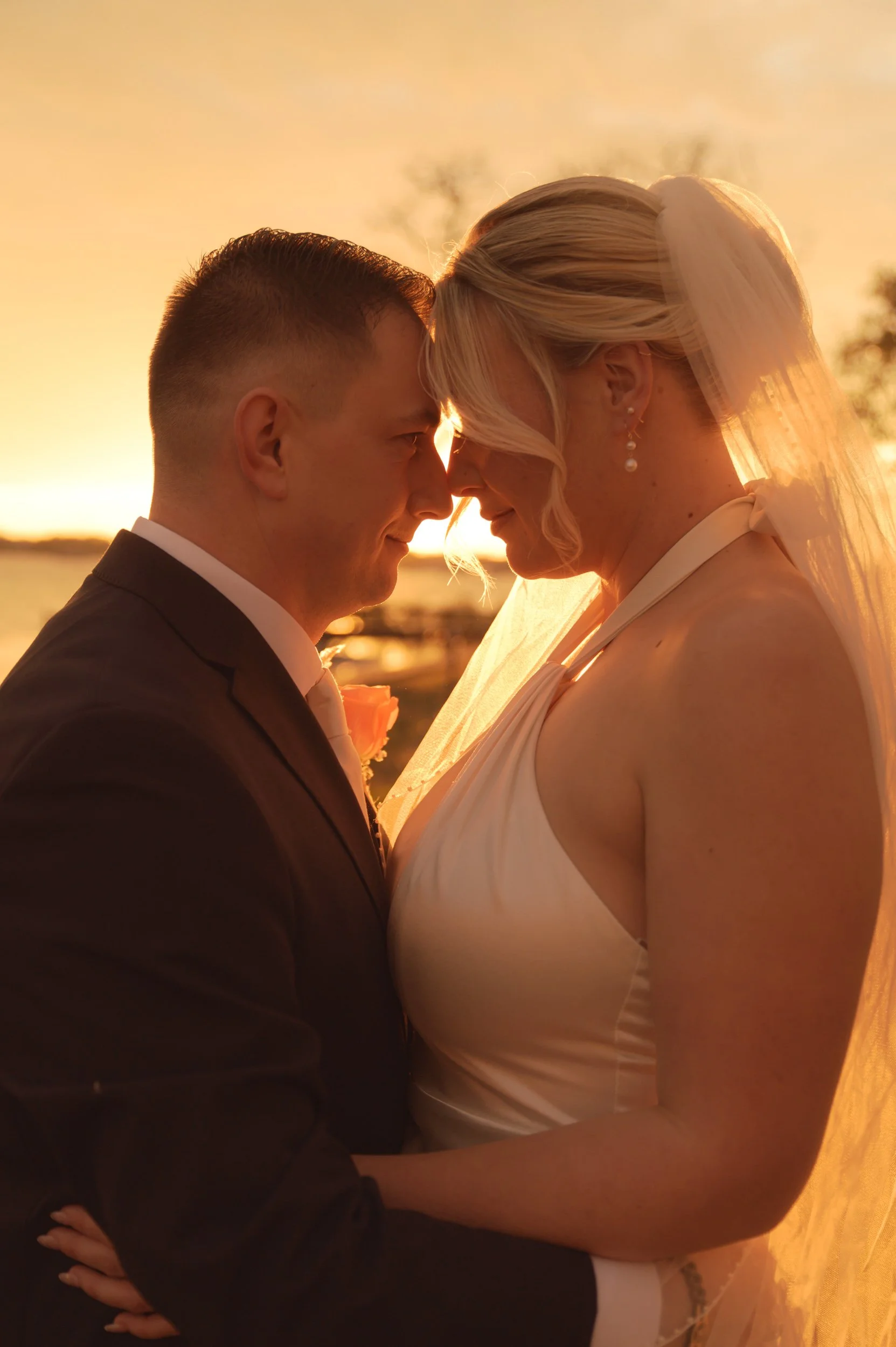 A bride and groom stand close together, with their foreheads touching during sunset, near a body of water, celebrating their wedding.