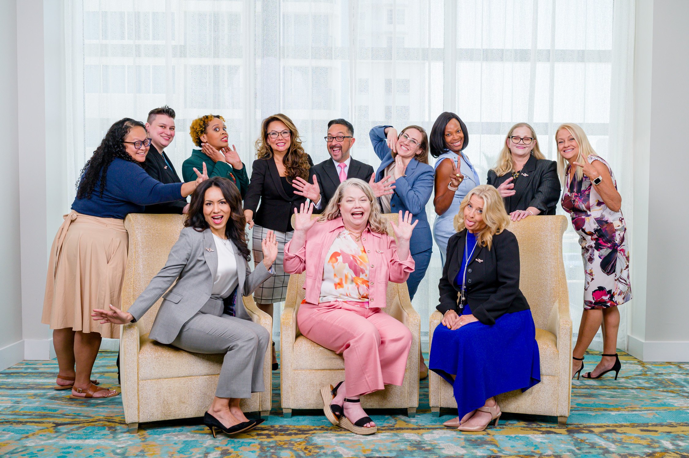 A diverse group of women and one man posing and smiling together in a bright room with a large window. Some are standing, some sitting, and they appear cheerful and friendly.