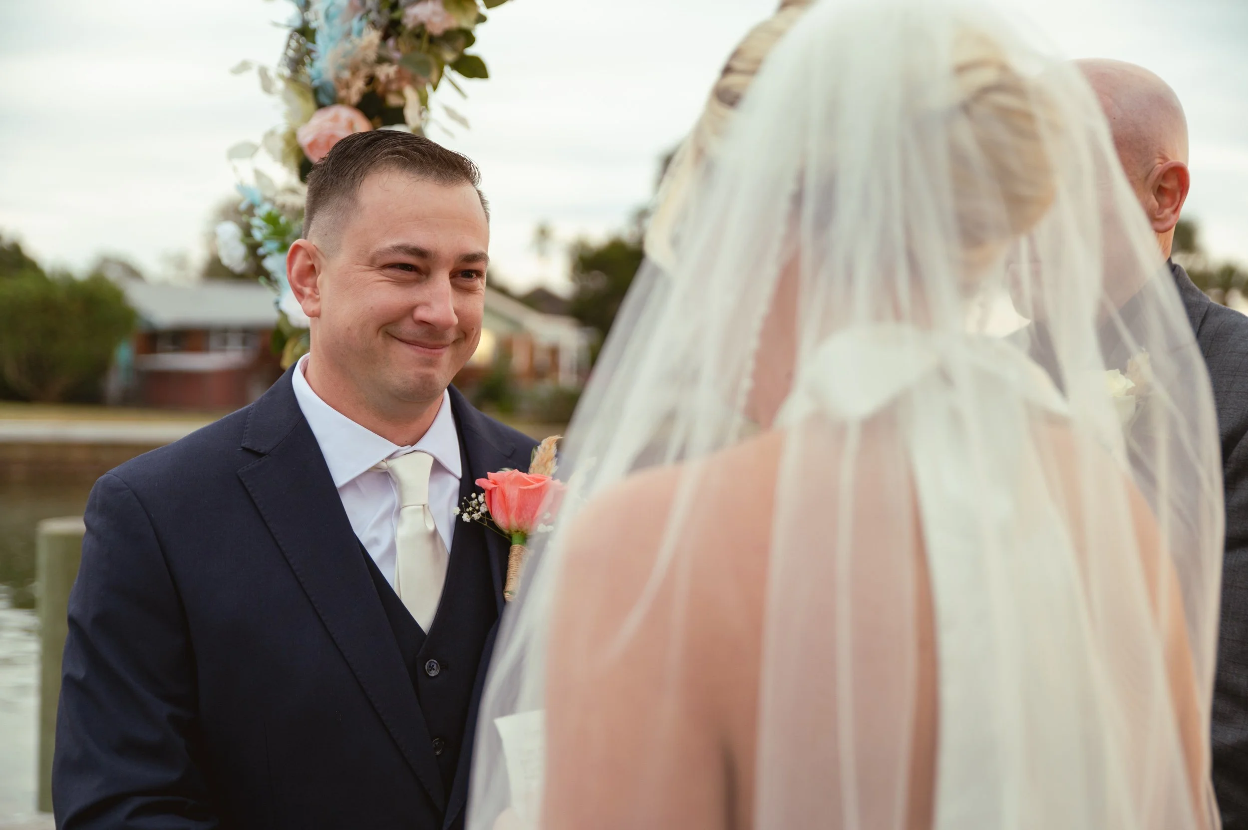 A groom in a dark suit with a white shirt and tie, smiling at a bride during an outdoor wedding ceremony near a body of water with houses in the background. The bride is wearing a veil and a strapless wedding dress, and holds a bouquet of flowers.