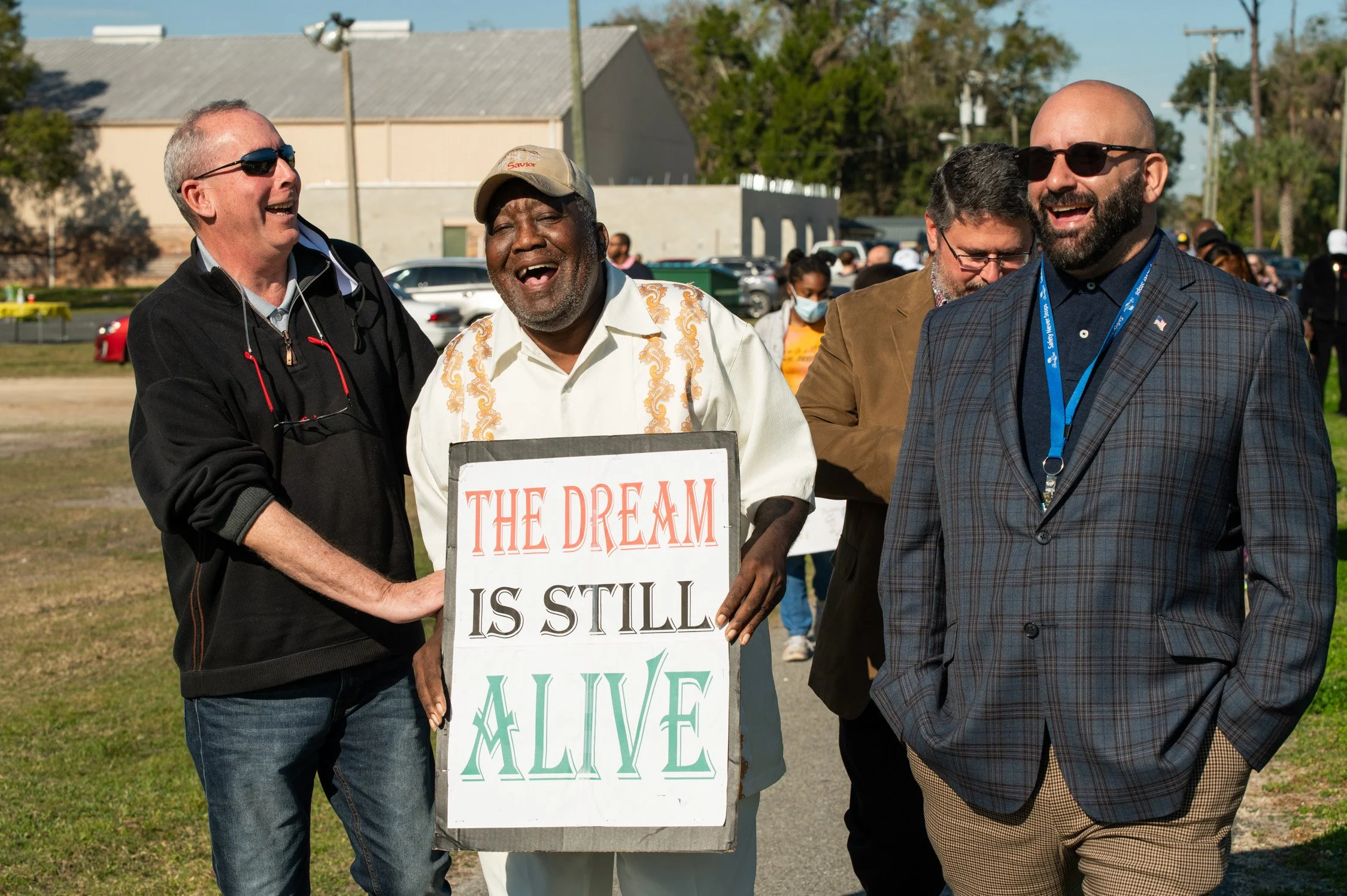 A group of people smiling and laughing outdoors, with one man holding a sign that reads "The Dream Is Still Alive."