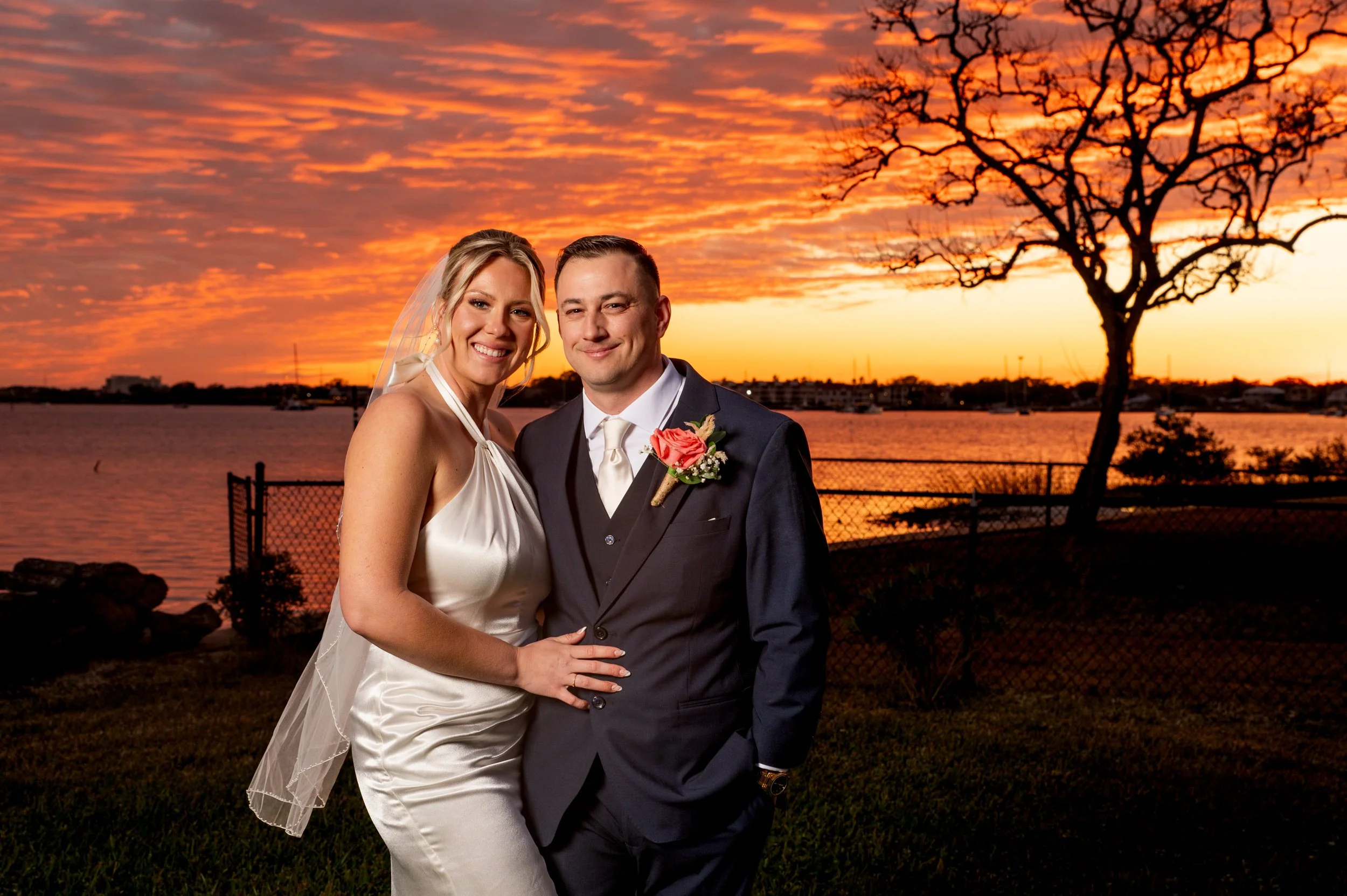 A happy bride and groom in wedding attire standing outdoors during sunset by a body of water, with a tree and a sunset sky in the background.