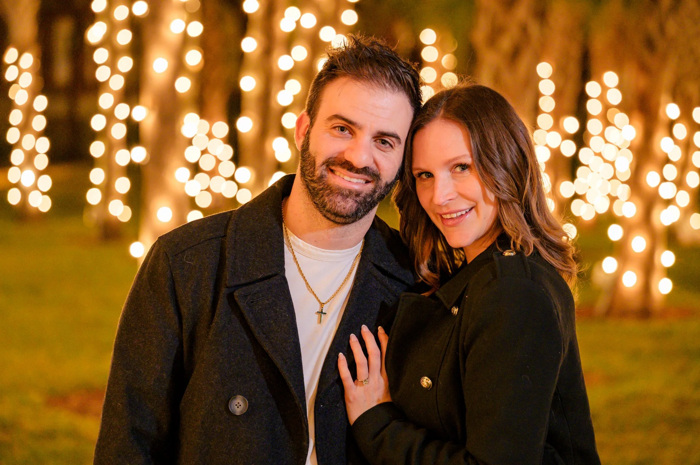 A smiling man and woman standing close together outdoors at night, with blurred string lights illuminating the background.