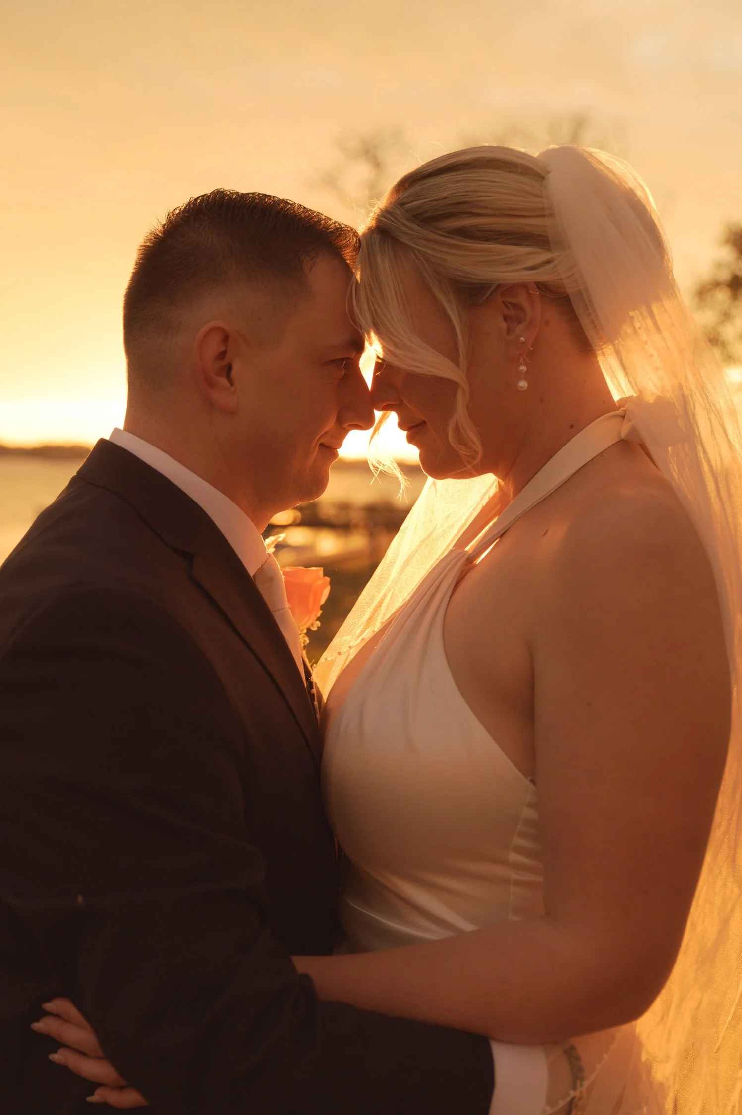 A newlywed couple stands close during sunset, with their foreheads touching and eyes closed. The groom wears a black suit and tie, and the bride wears a white wedding dress with a veil. The background shows a body of water and a sunset sky.