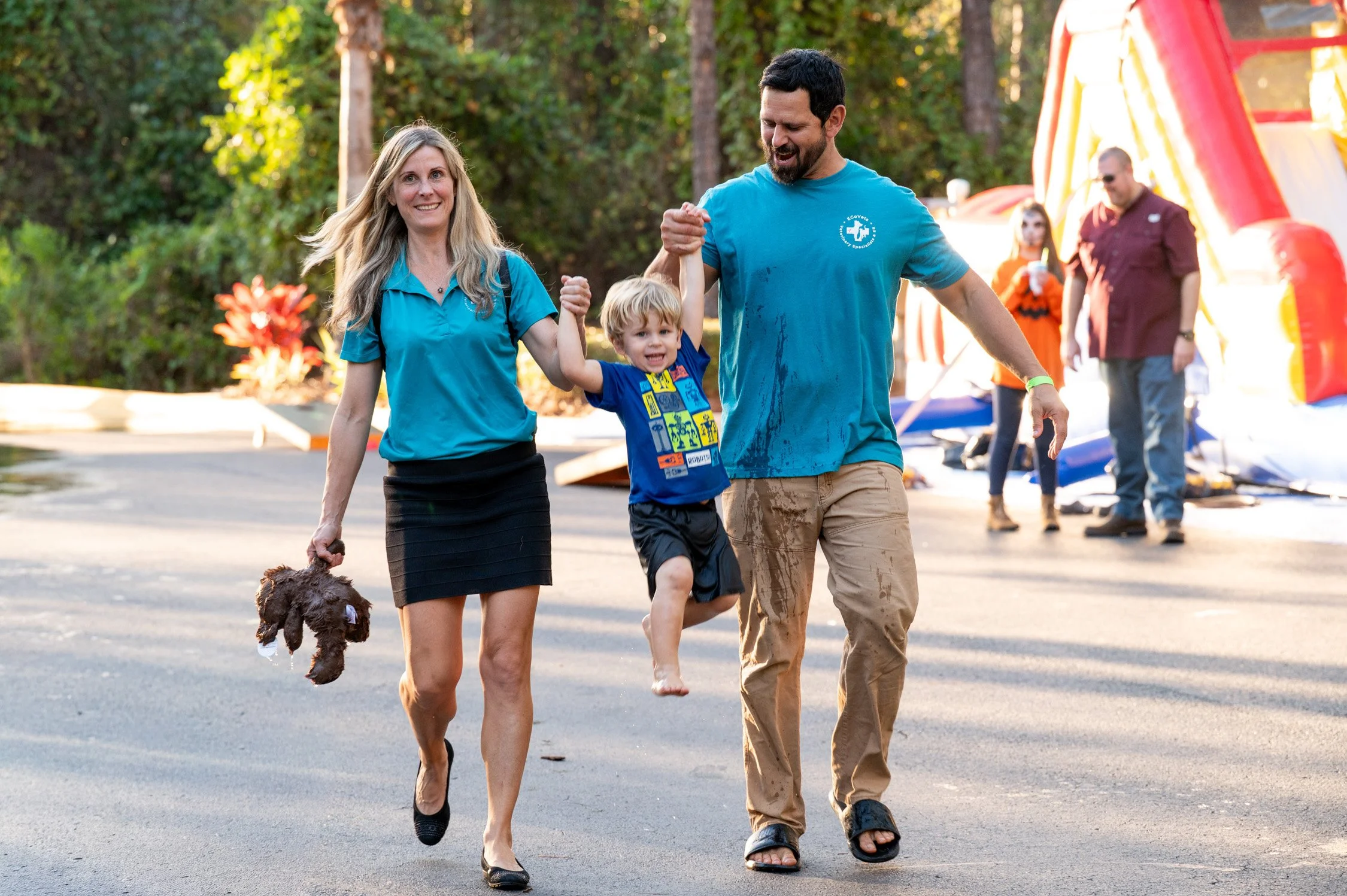 Family celebrating outdoors, young boy being lifted by father, mother holding a stuffed animal, in a park or camping area with trees and inflatable water slides in background.