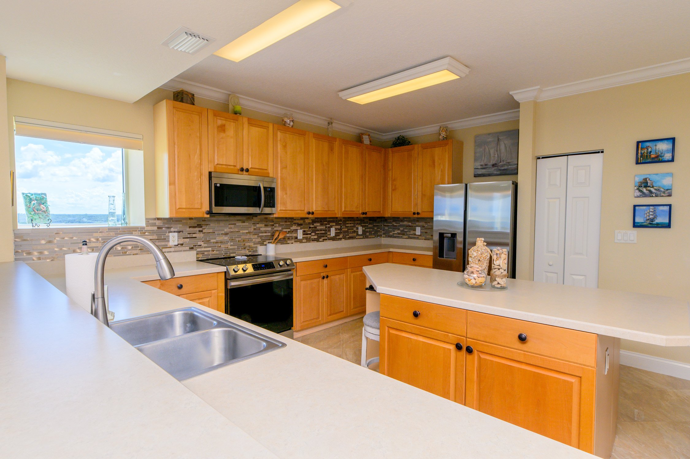 Bright kitchen with wooden cabinets, a double sink, beige countertops, stainless steel appliances, and nautical artwork on the walls.
