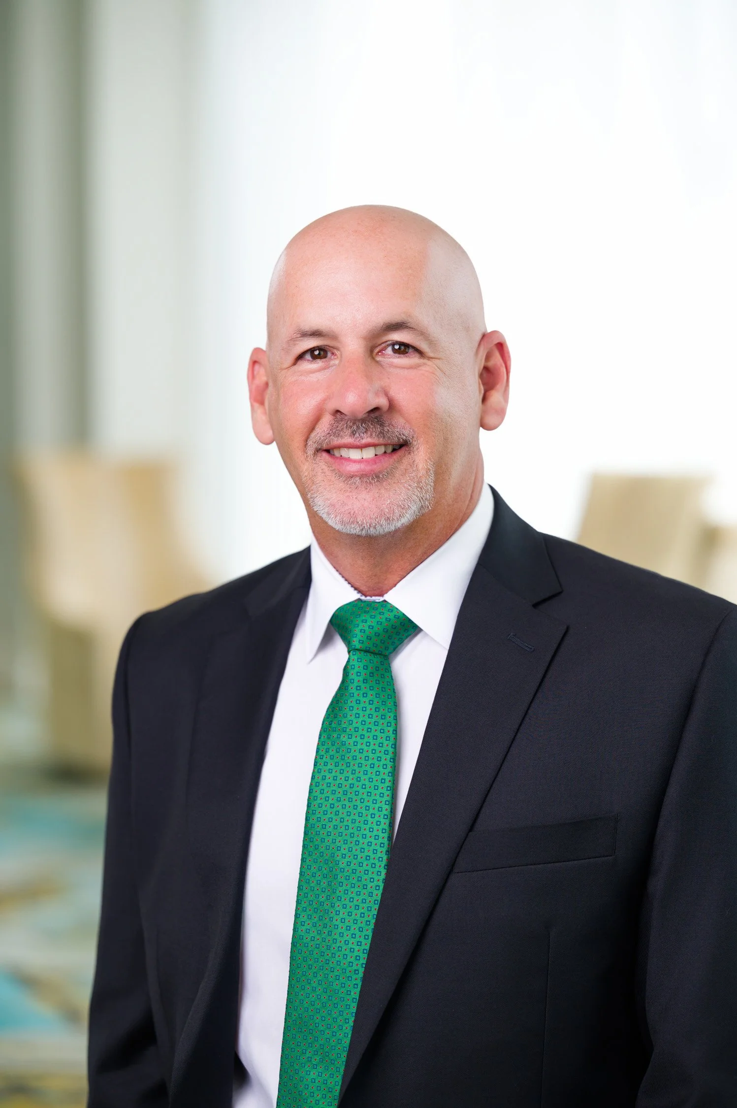 A portrait of a middle-aged, bald man with a goatee, wearing a black suit, white shirt, and green tie, smiling in a professional setting.