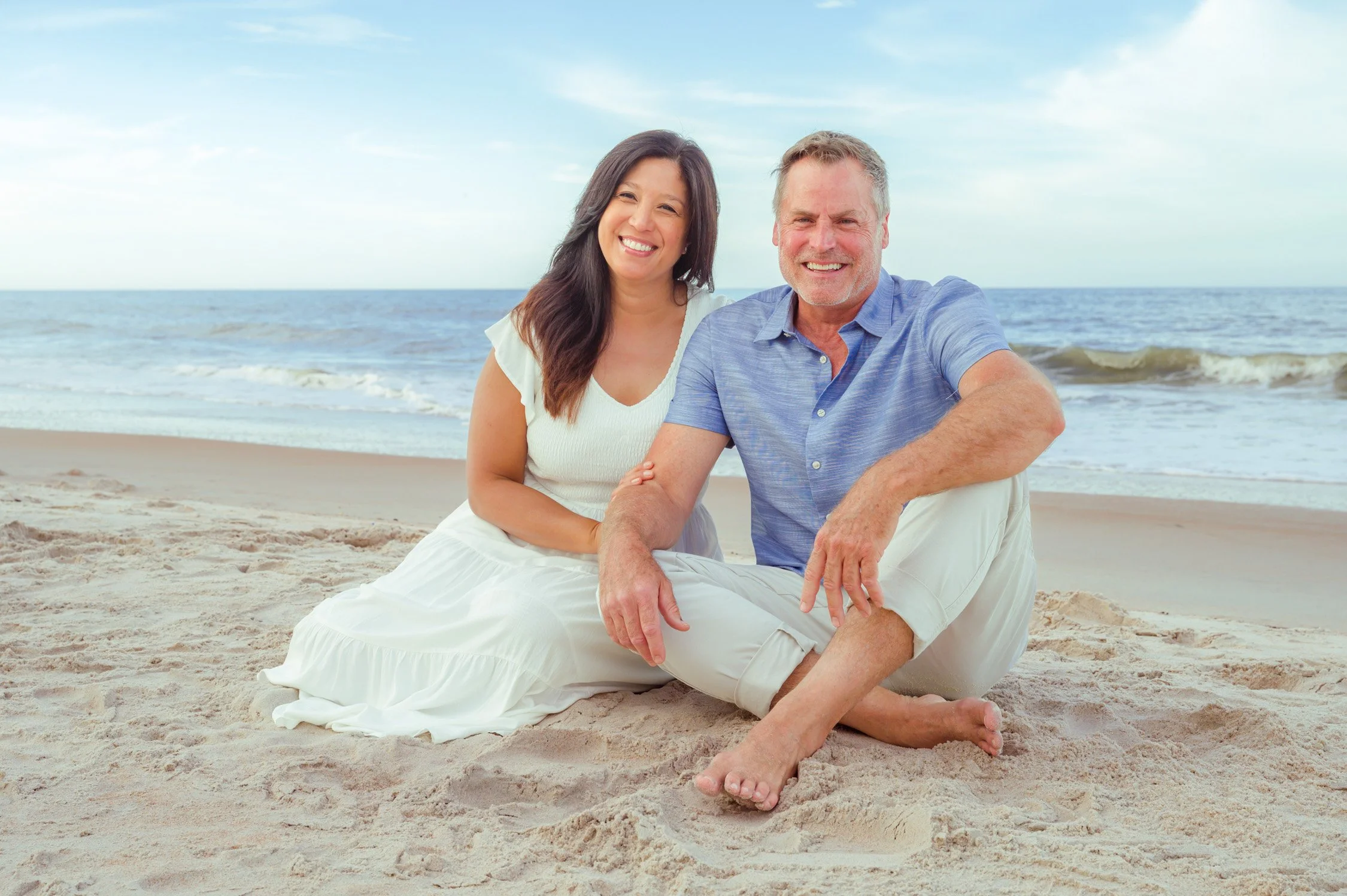 A smiling couple sitting on the beach with the ocean in the background.