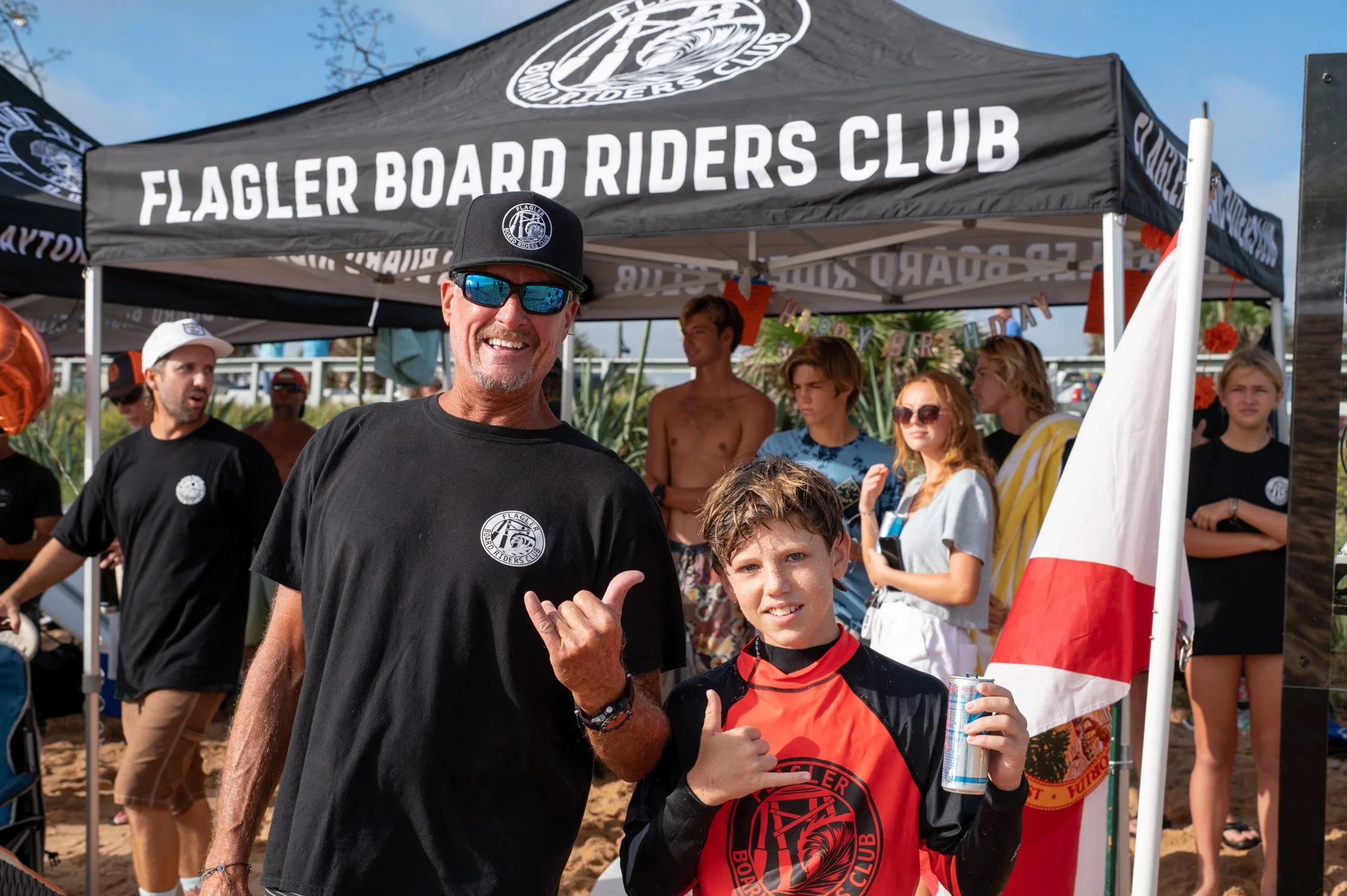 People celebrating at a Flagler Board Riders Club event on the beach, with a man and a boy in the foreground, a canopy tent, and a group of young people in the background.