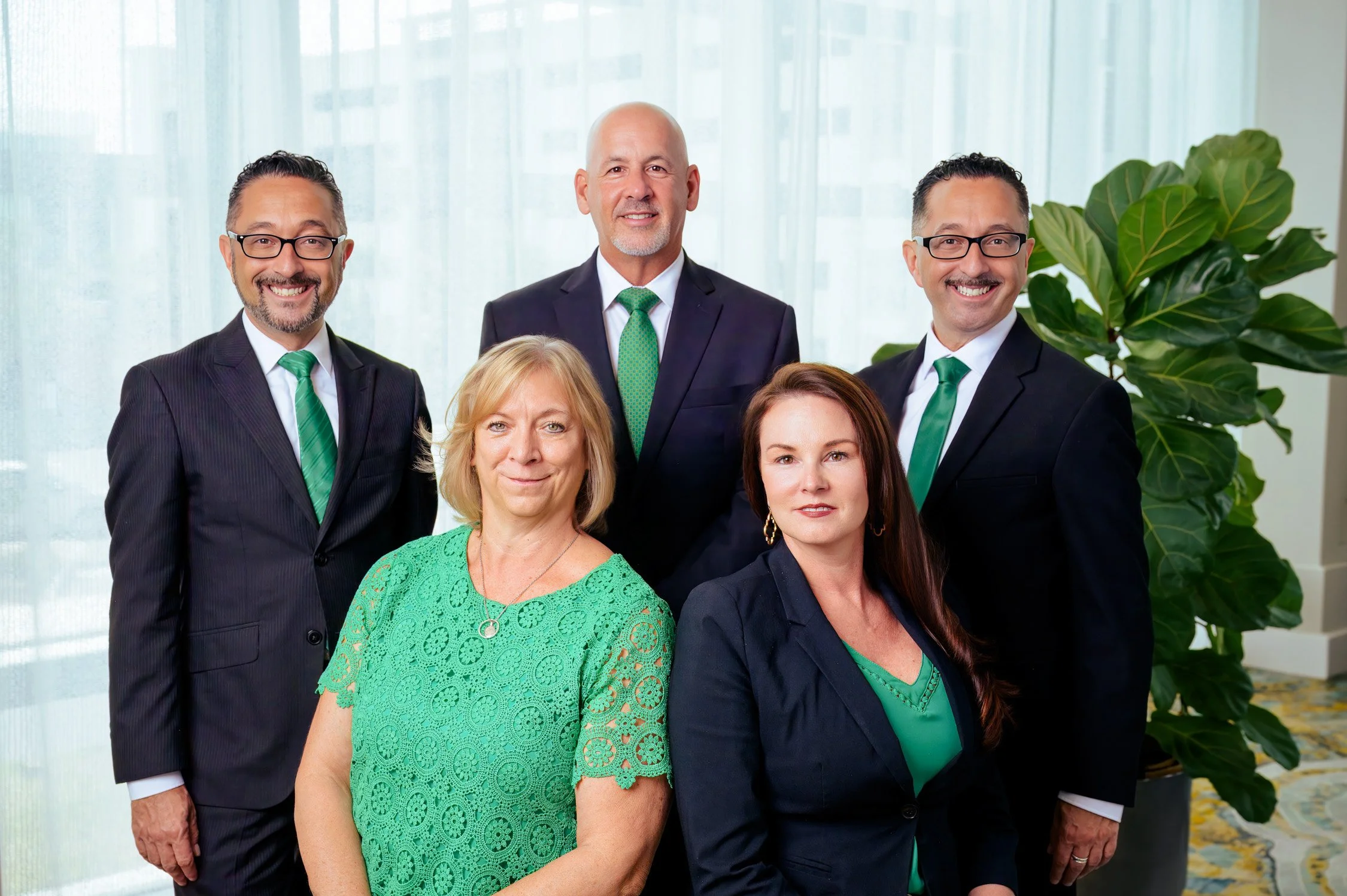 Group of six diverse professionals in business attire posing for a photo indoors with large window and green plant in the background.