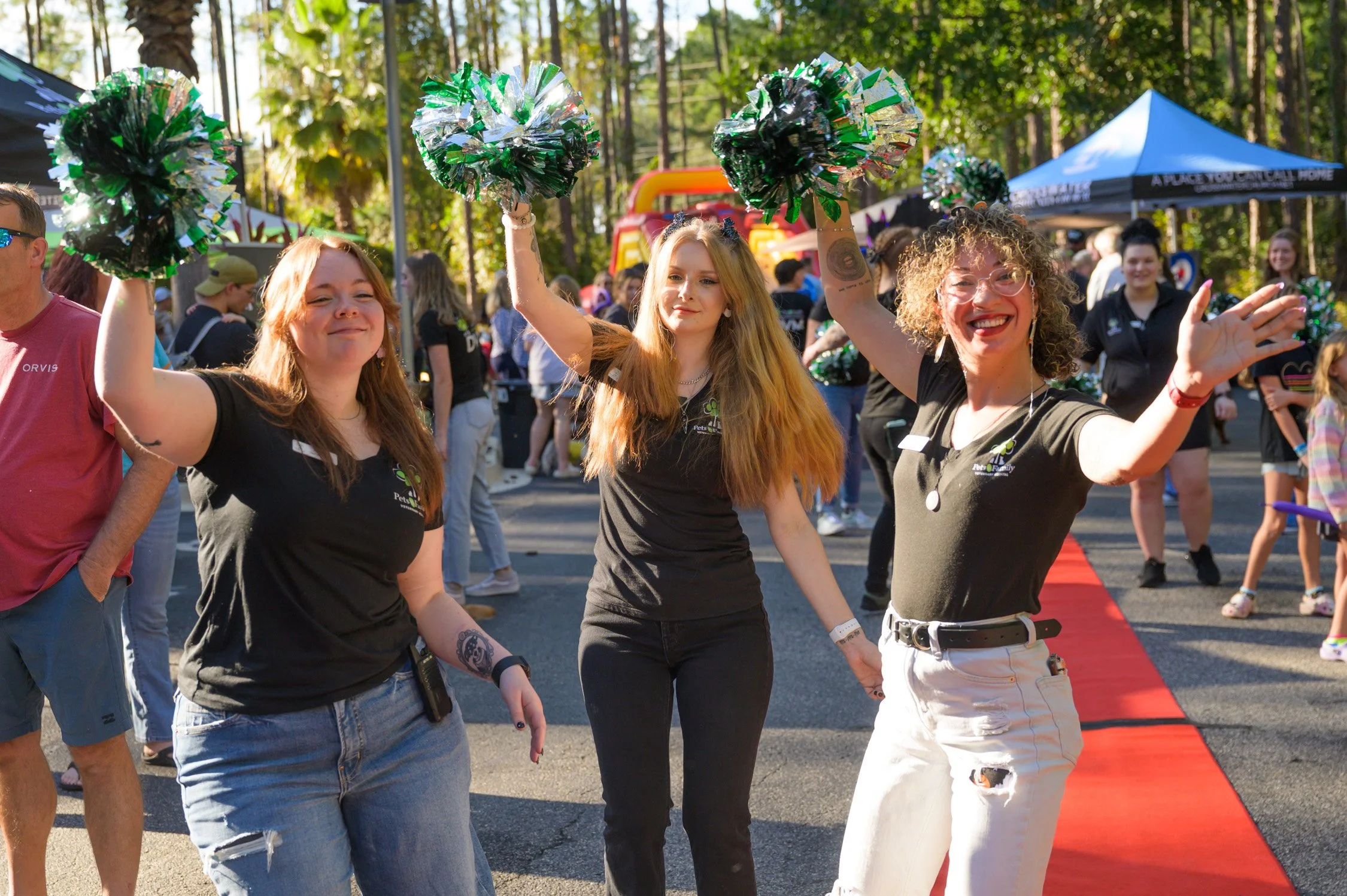 Three women cheerfully holding green and silver pom-poms at an outdoor event, with other attendees and a red carpet in the background.