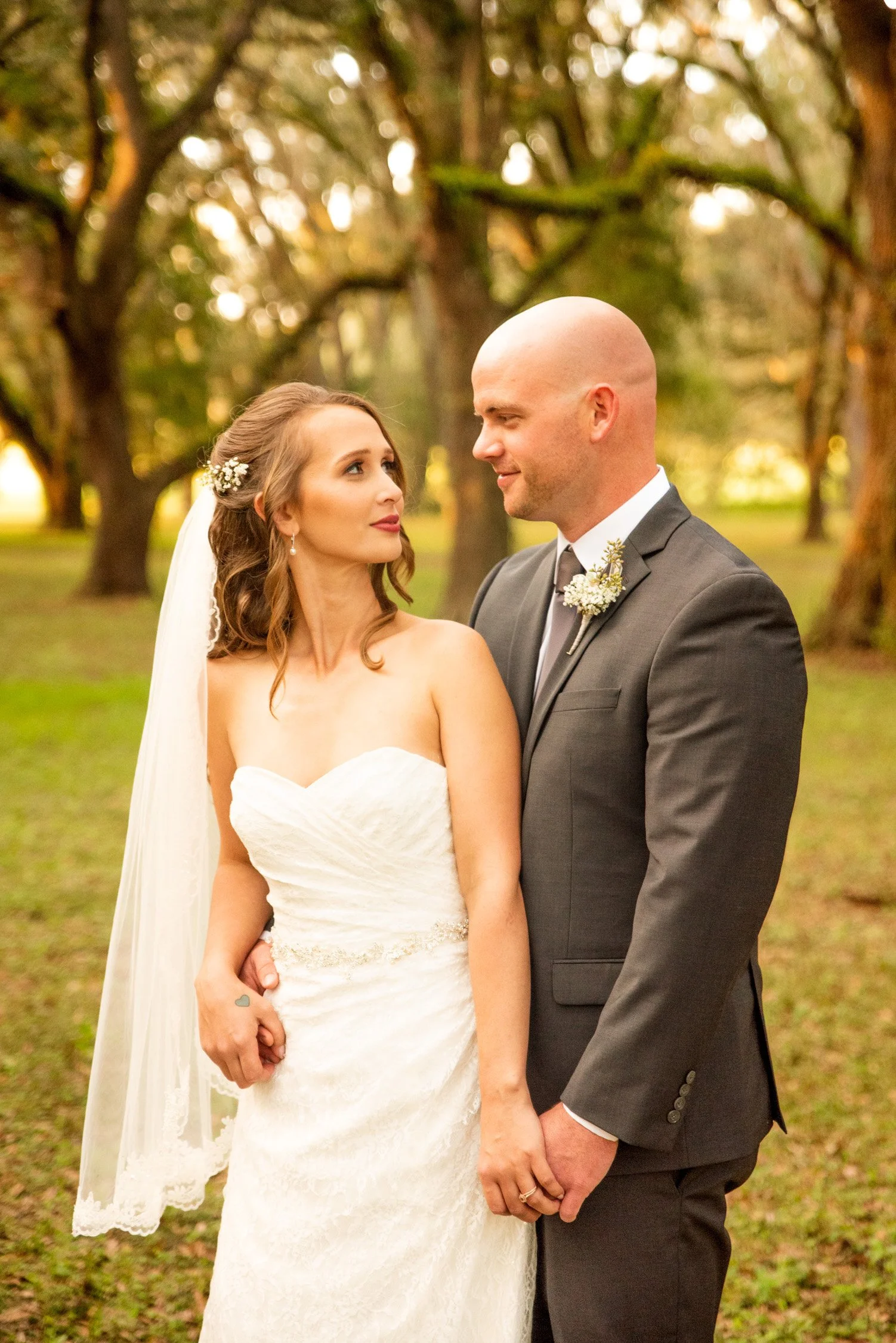 Bride and groom holding hands in a park with trees, looking at each other.