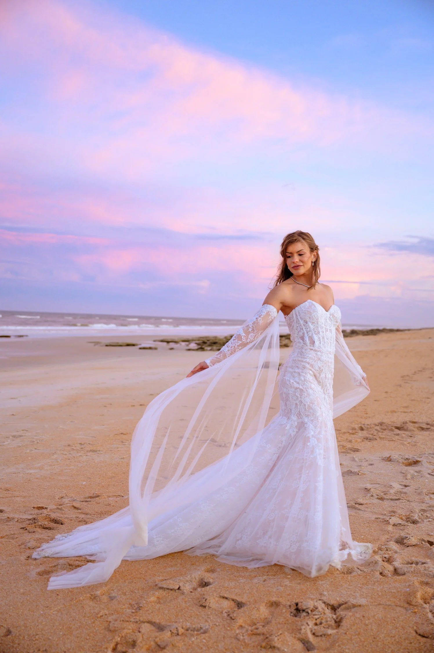 A woman in a white lace wedding dress stands on a sandy beach at sunset, looking contemplative with a pastel sky in the background.