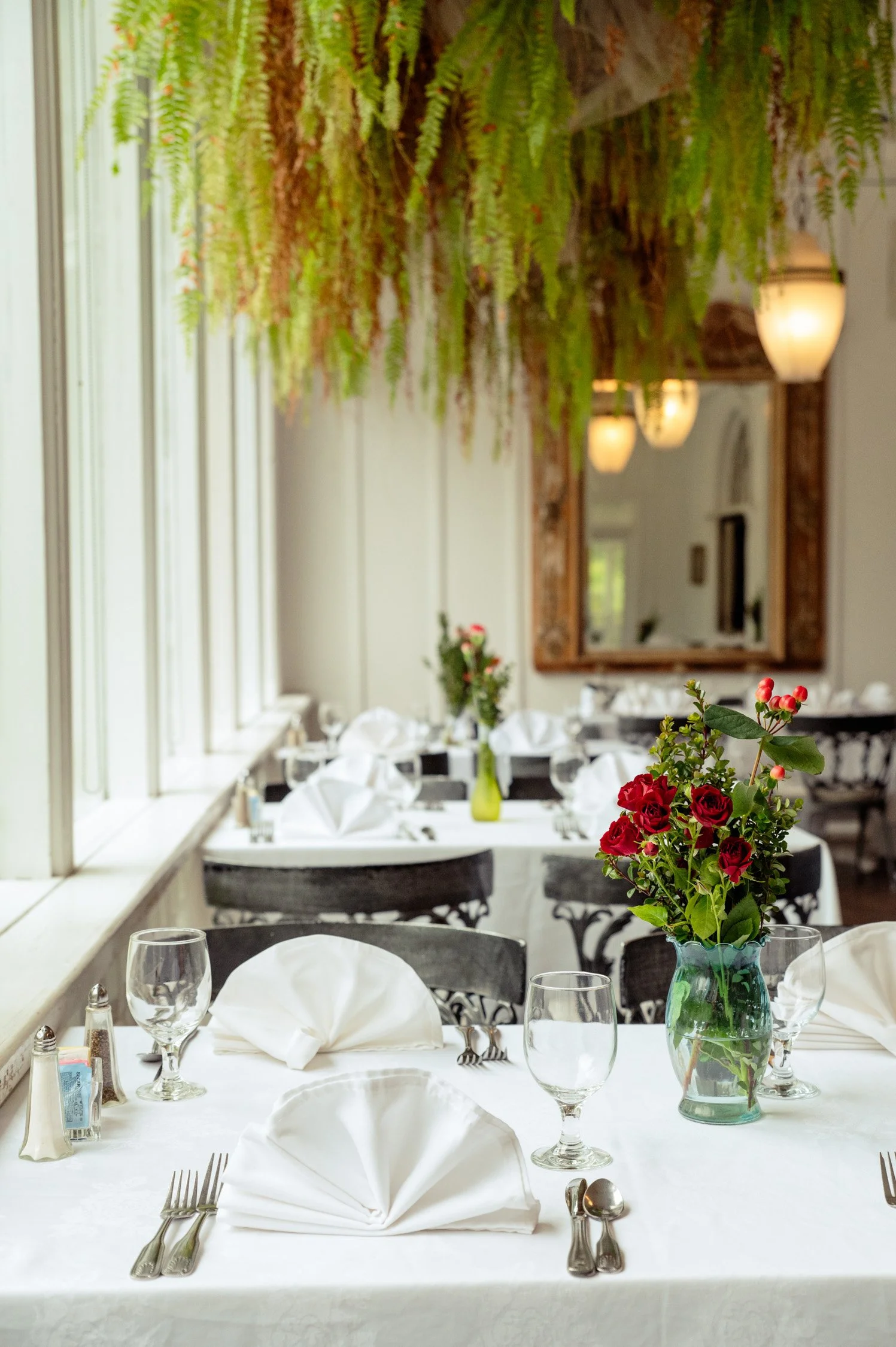 A restaurant table set for dining with white tablecloth, white folded napkins, glassware, silverware, and a floral centerpiece with red and pink flowers in a glass vase, in a bright room with large windows, hanging green plants, and warm lighting.