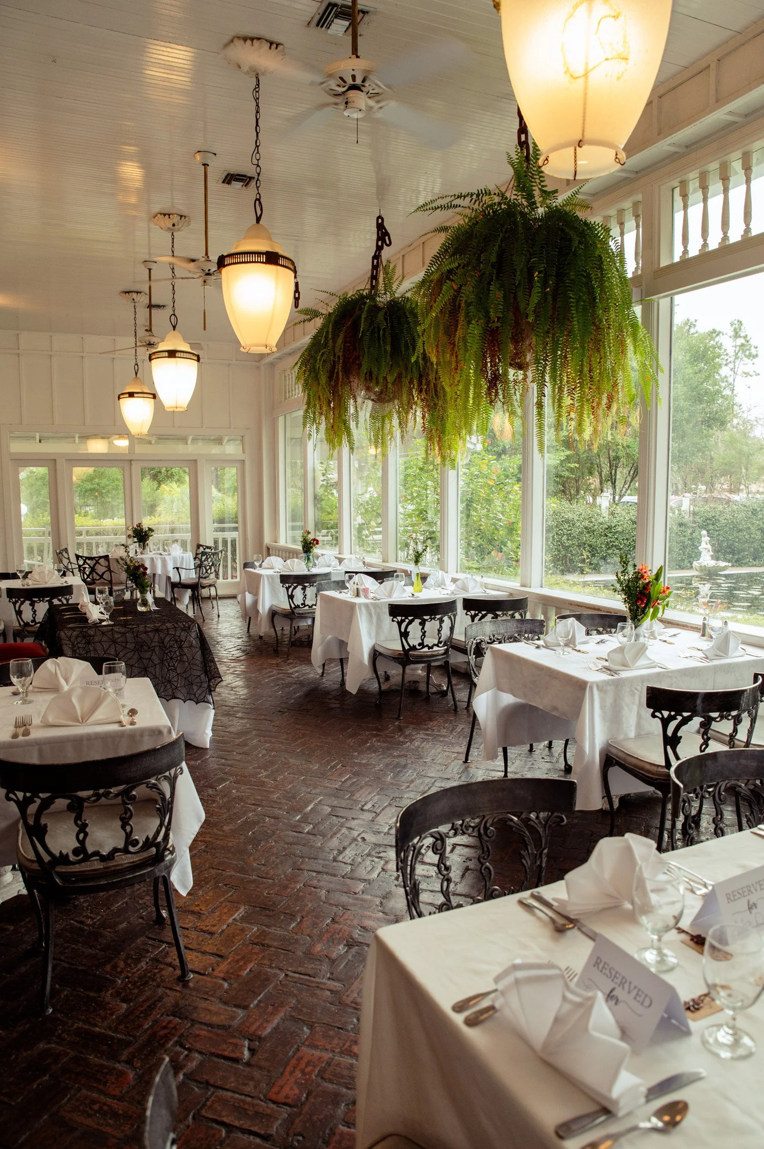 Elegant restaurant dining room with white tablecloths, napkin art, and reserved signs on tables. Large hanging ferns and warm ambient lighting. Windows with green outdoor scenery.