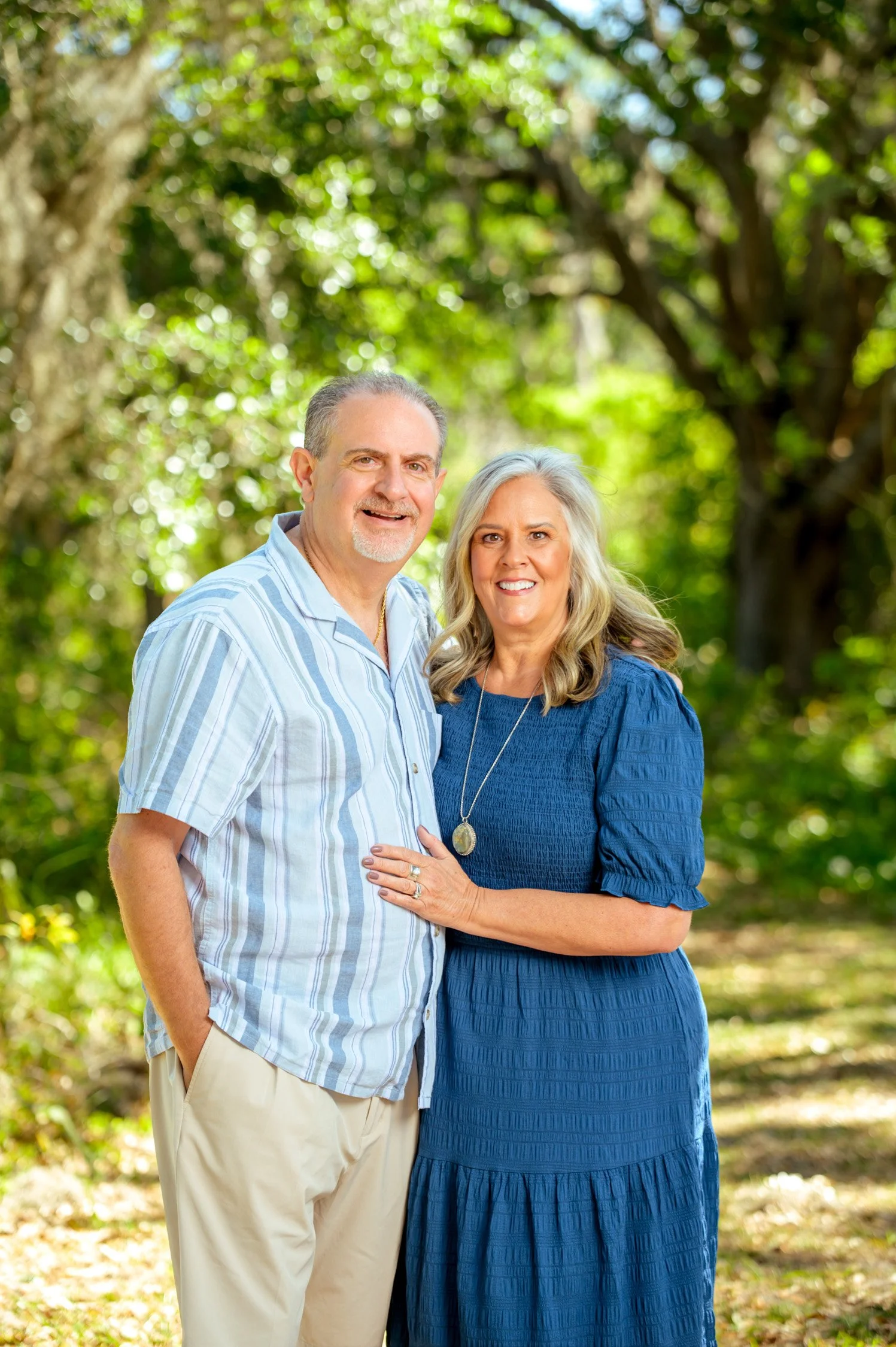 A smiling middle-aged couple standing outdoors in a wooded area during daytime. The man has short gray hair and a beard, wearing a light blue striped shirt and beige pants. The woman has long blonde hair, wearing a blue dress with a pendant necklace,