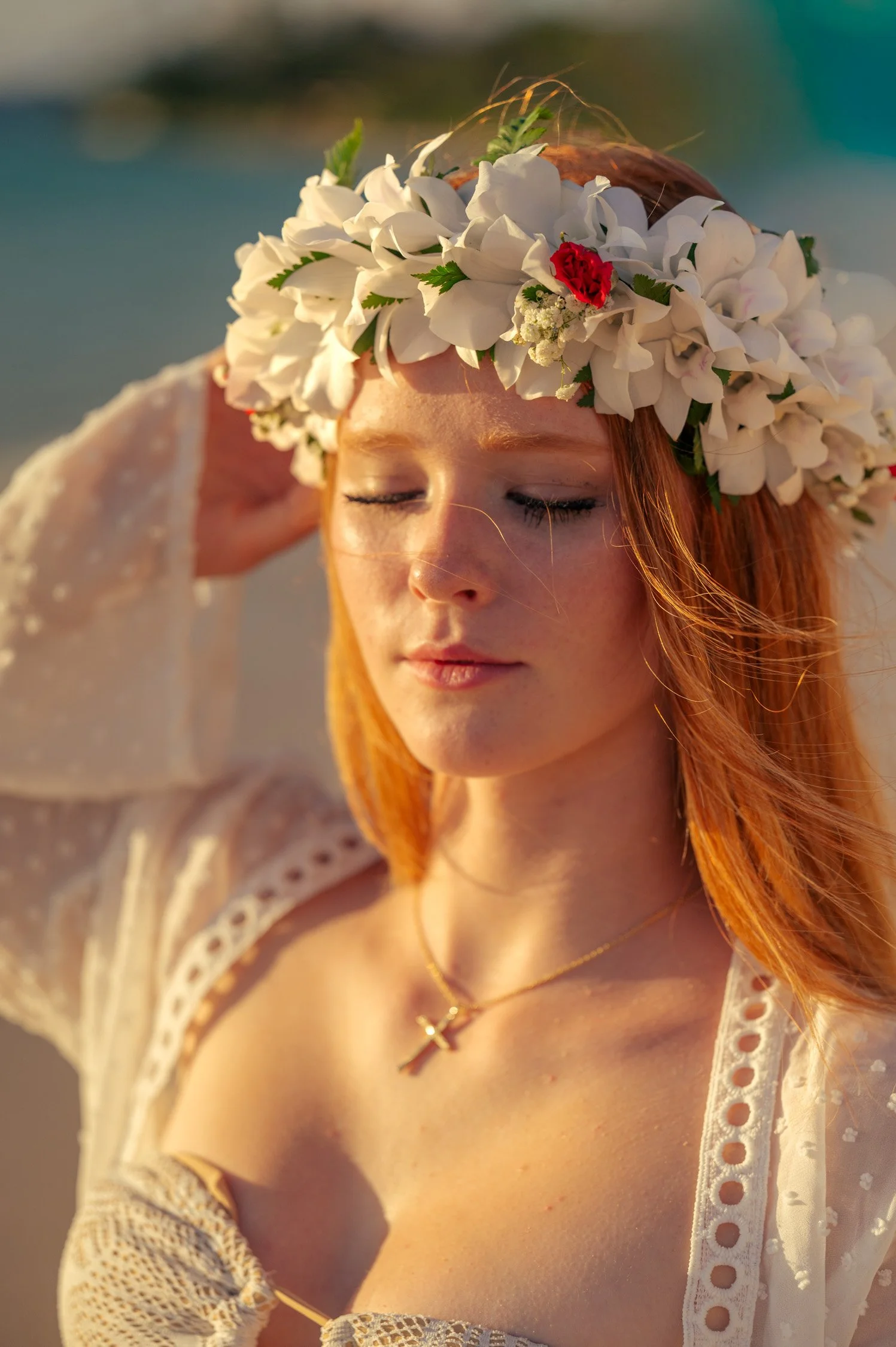A young woman with red hair and closed eyes, wearing a floral crown with white flowers and a small red flower, standing outdoors in warm sunlight.