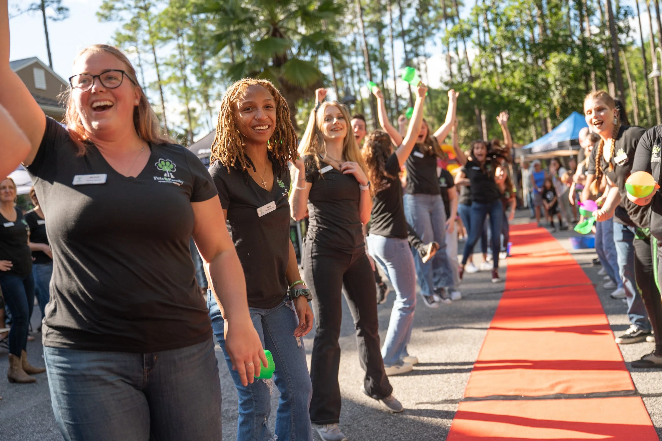 Group of women wearing black shirts participating in outdoor event, some holding water balloons, on a red carpet with trees and tents in the background.