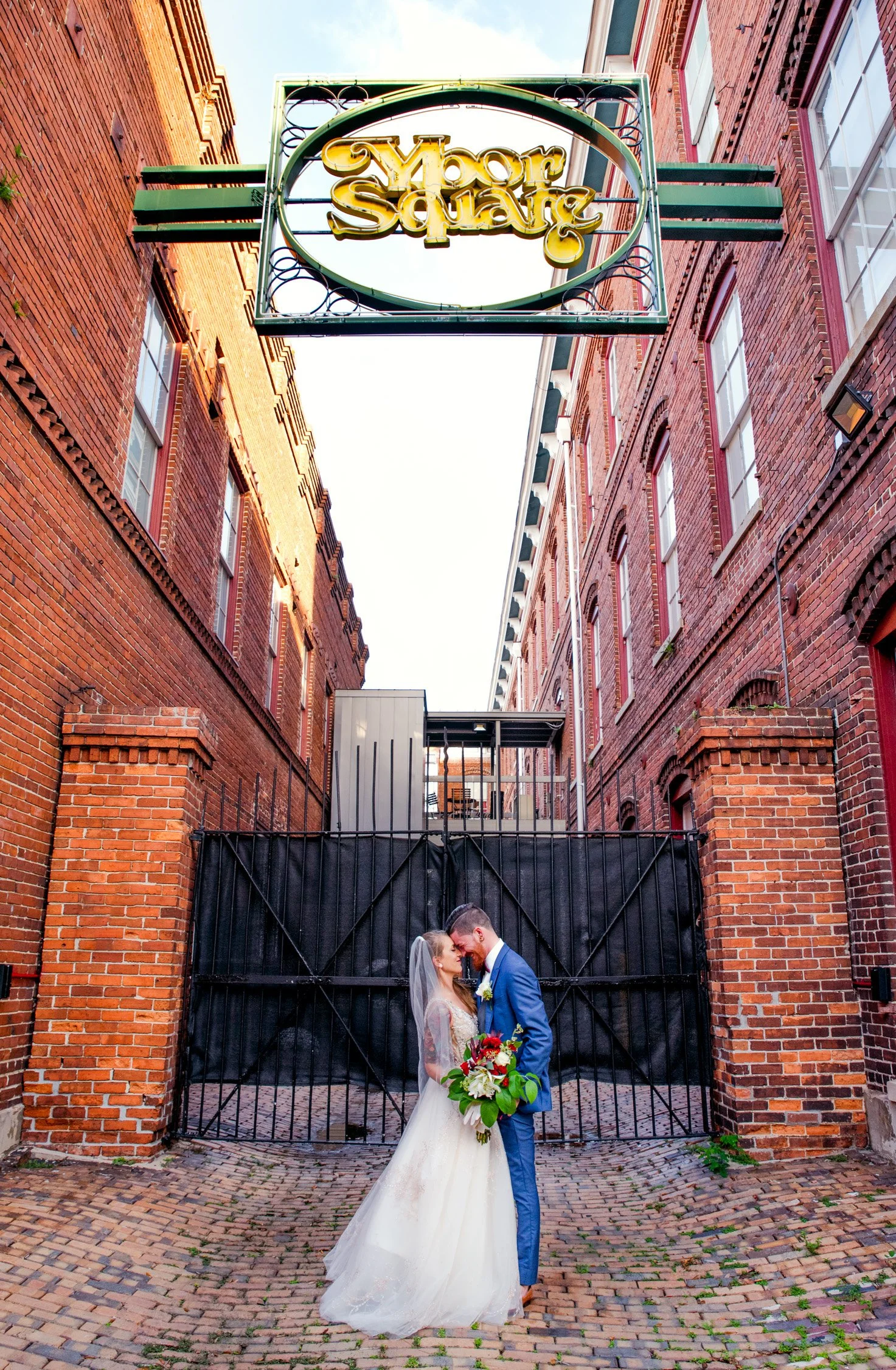 A bride and groom standing close and touching foreheads in front of a black gate between red brick buildings, with a sign overhead that reads "Moore Square." The bride is wearing a white wedding dress and holding a bouquet of flowers, while the groom