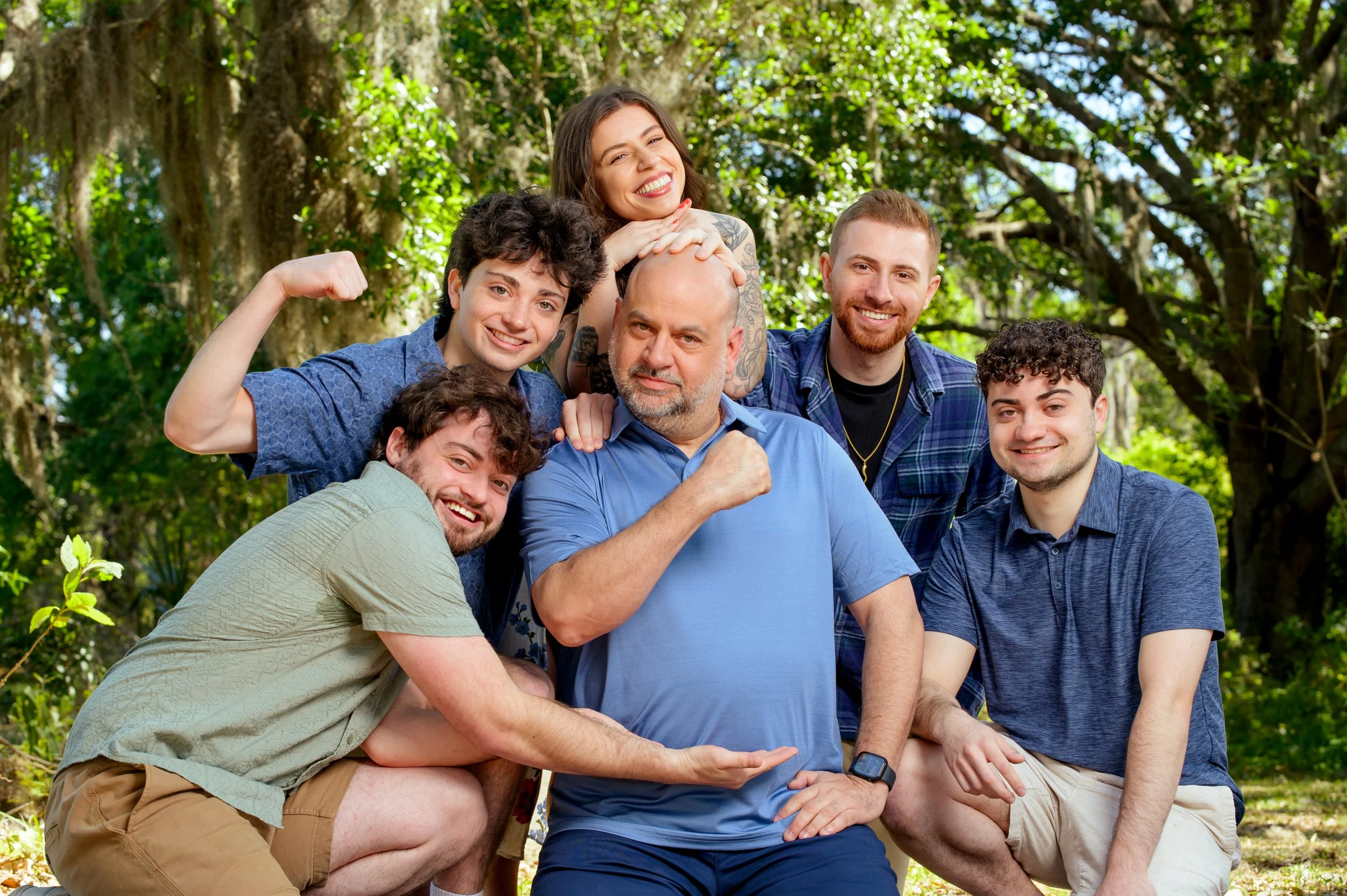 Group of six people outdoors in a wooded area, smiling and posing together, with some flexing muscles and one person making a fist.