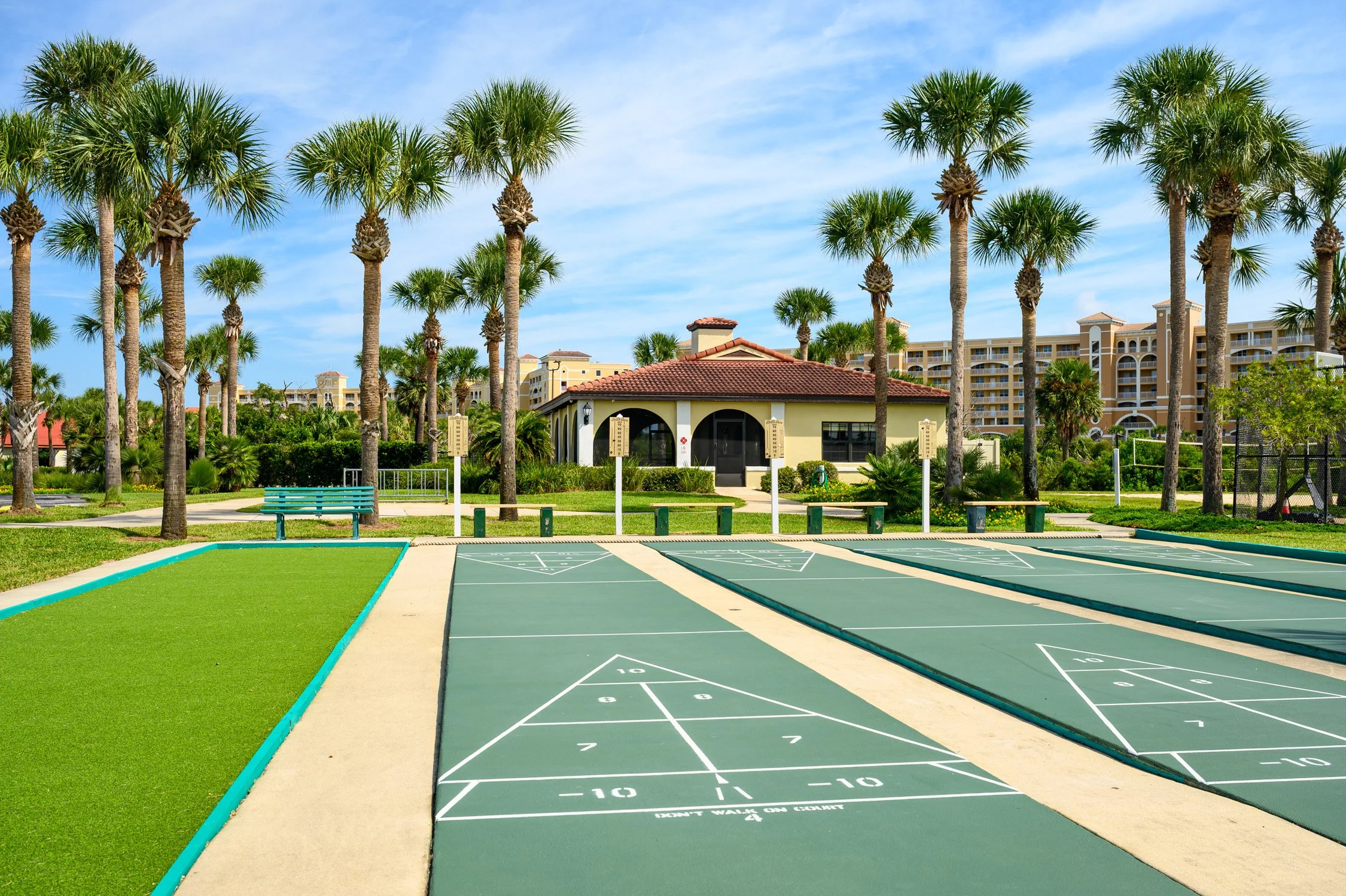 An outdoor shuffleboard court with multiple courts, surrounded by palm trees, a grassy area, and a building in the background under a blue sky.