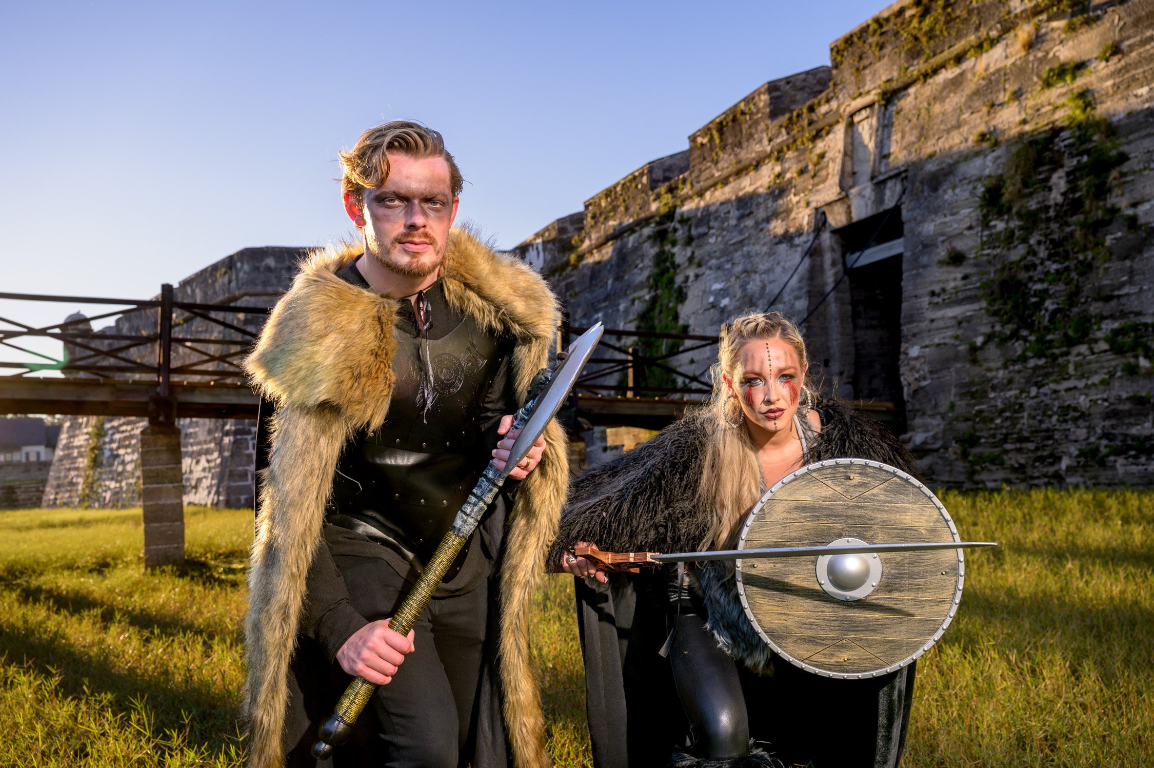 Two people dressed as fantasy warriors outdoors, with a stone fortress in the background. The person on the left is a man wearing a fur cape and black armor, holding a spear. The person on the right is a woman dressed in a black cloak, with face pain