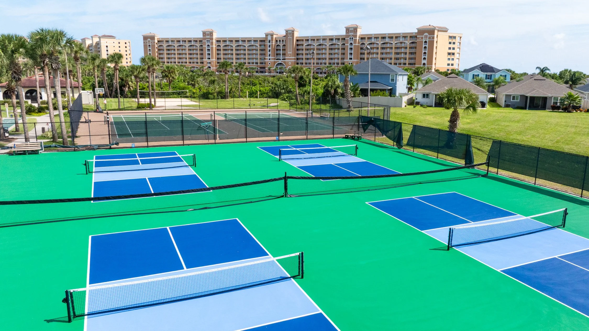 Multiple blue and green tennis courts with nets surrounded by a chain-link fence, with residential houses and a large apartment building in the background, under a partly cloudy sky.