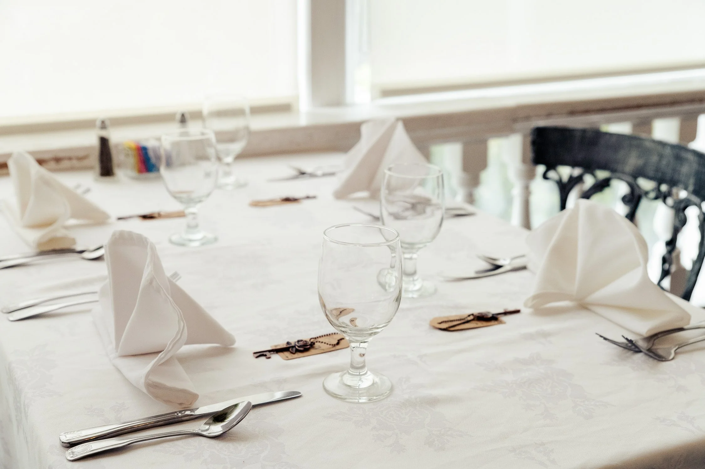 Formal dining table set with white cloth, folded napkins, wine glasses, and silverware, near a window with natural light.