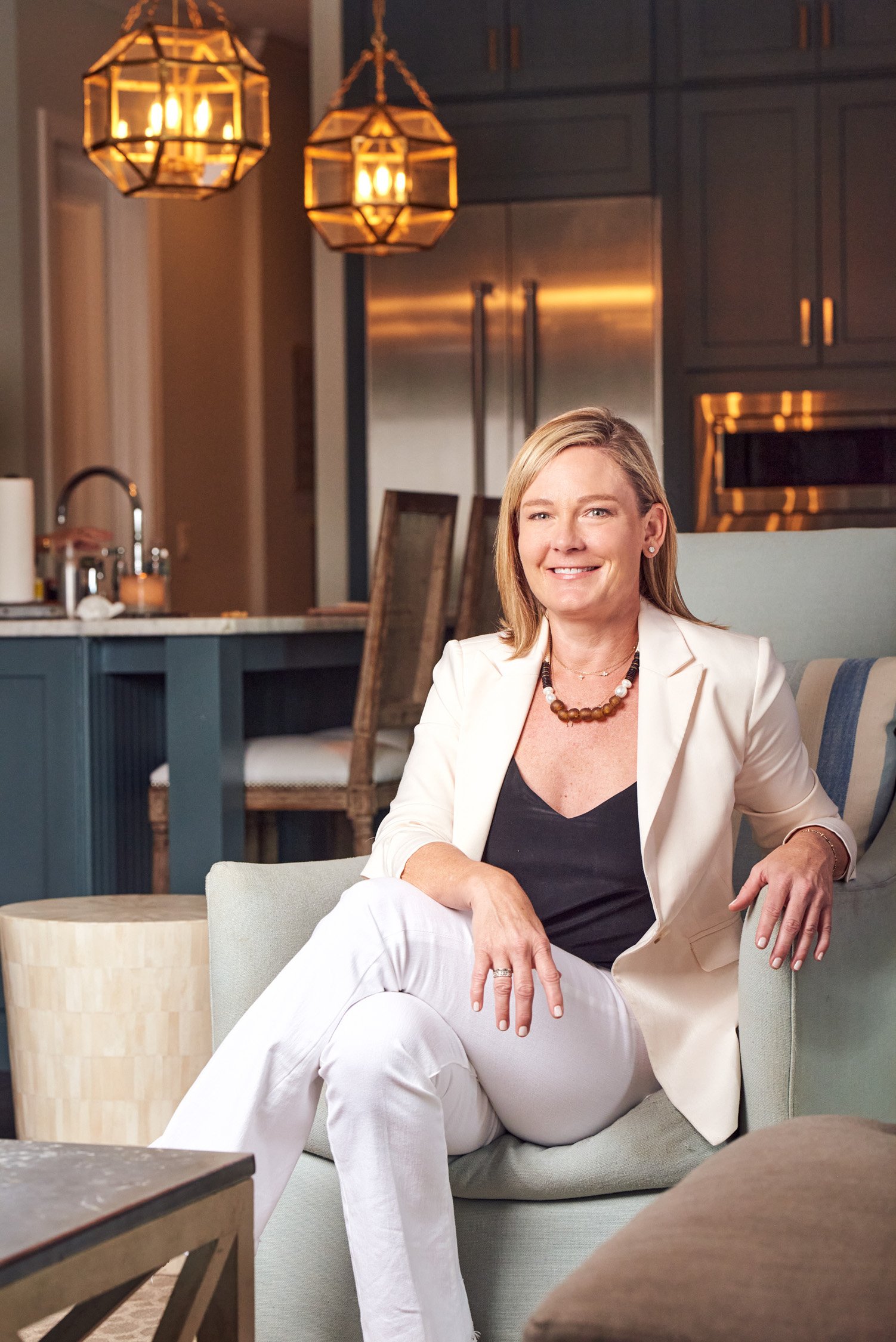 A woman sitting on a light-colored sofa in a modern kitchen, smiling at the camera. She has blonde hair and is wearing a white blazer, black top, white pants, and a beaded necklace. The kitchen features dark cabinets, a stainless steel fridge, and ha
