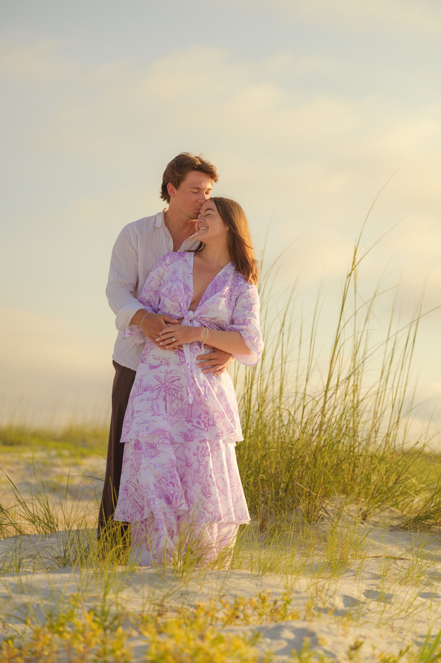 A couple standing on a sandy beach with tall grass, embracing and smiling at each other during sunset.